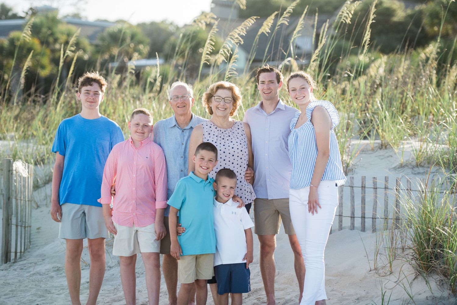 Large family group poses for casual beach photos with coastal vegetation and fencing.