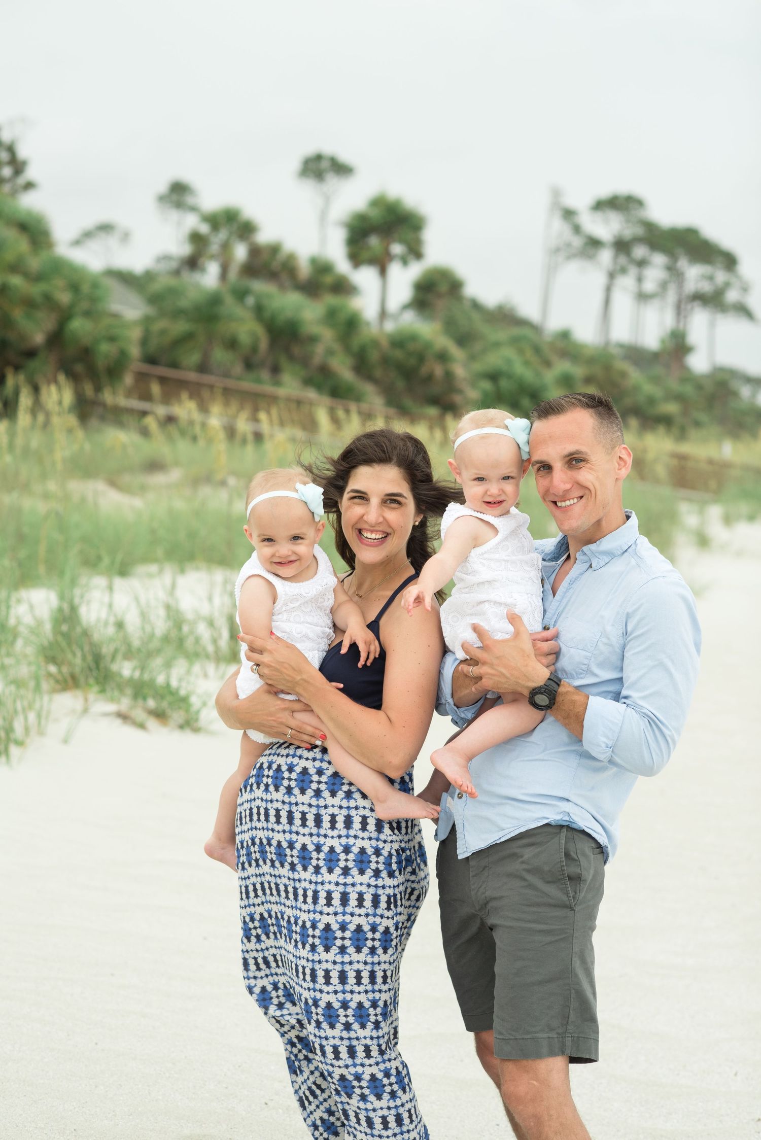 Happy family photo session with twin babies posing on a white sand beach with palm trees in the background.