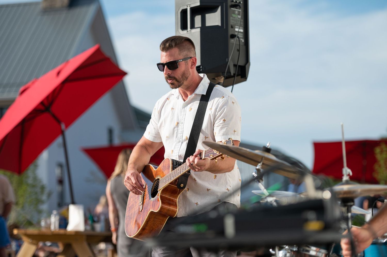 Musician performs on stage with acoustic guitar under red umbrellas during outdoor summer concert.