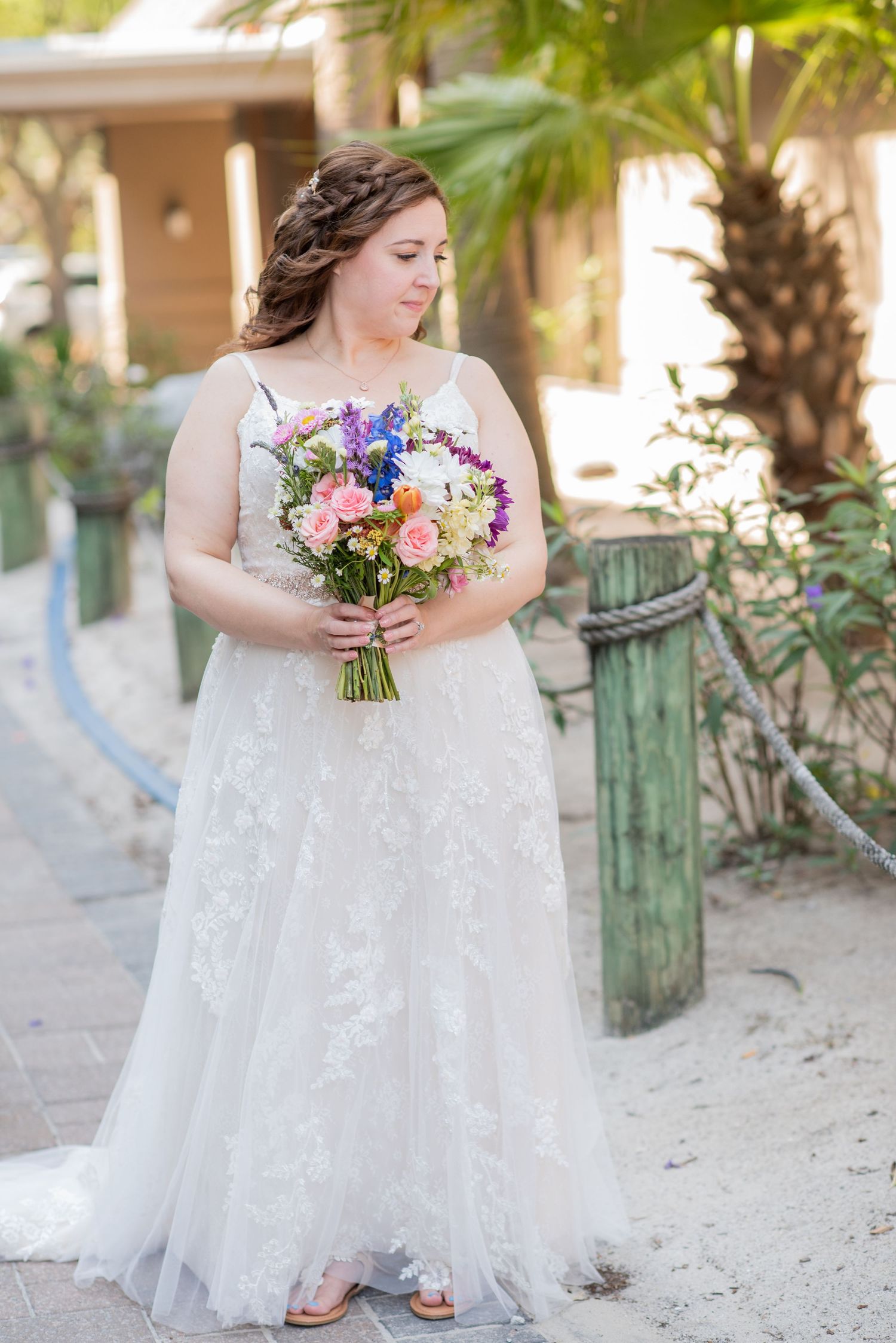 A bride in a white lace gown holds a colorful bouquet while standing on a sandy beach path with palm trees.