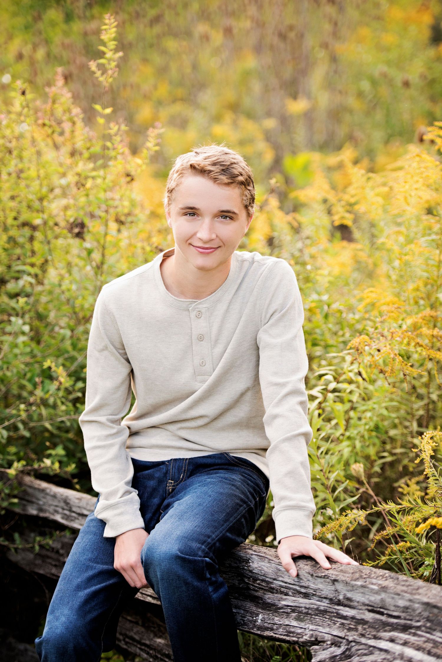 Portrait of a young person in a beige henley sweater sitting on a rustic log with yellow wildflowers in background.