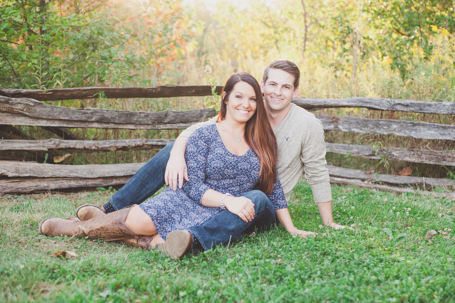 Couple lounges together on grass near rustic wooden fence during sunset photoshoot.
