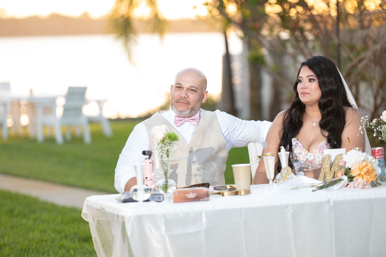 Bride and groom seated at sweetheart table during outdoor waterfront wedding reception at golden hour.