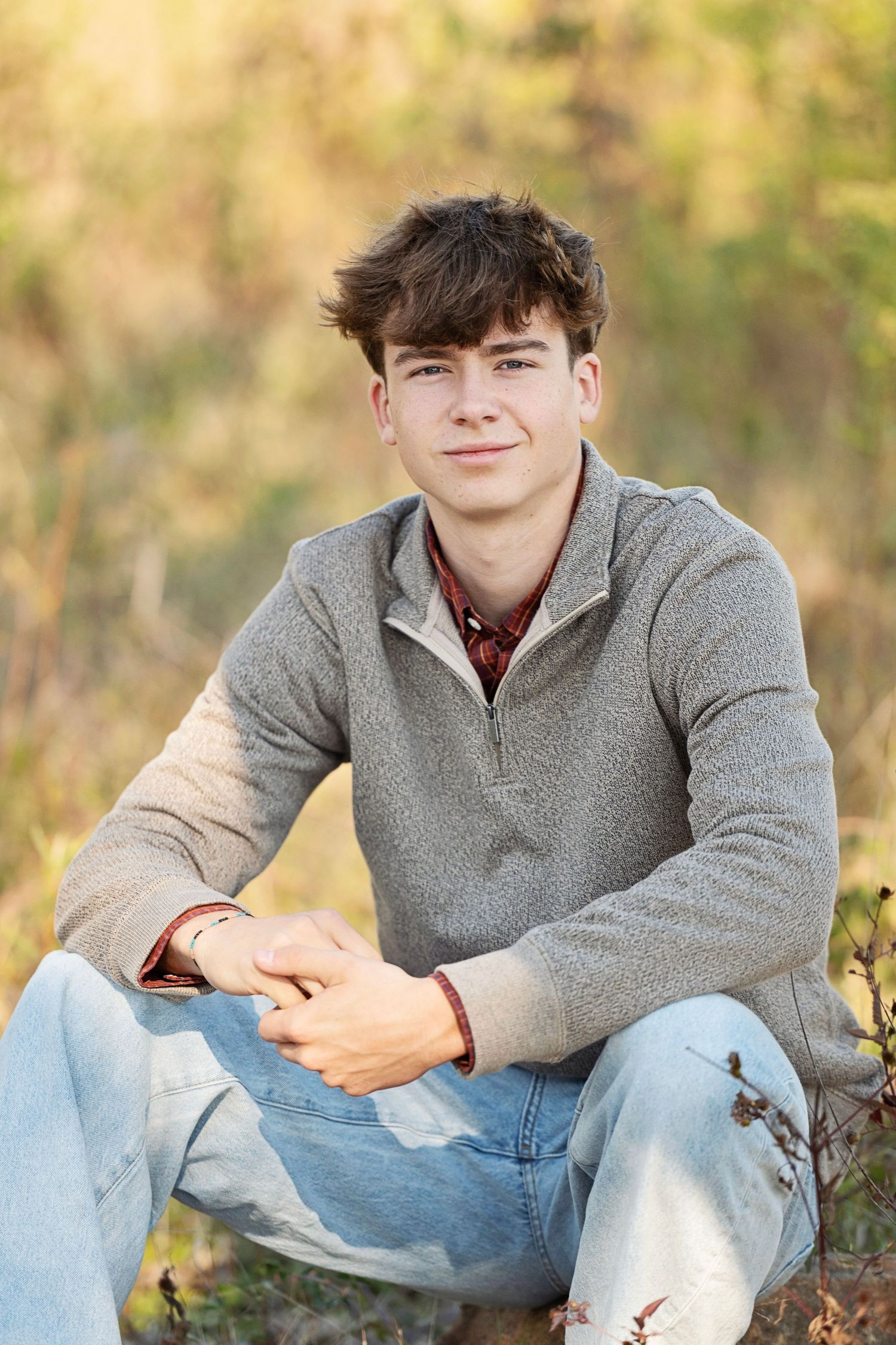 Young man in gray pullover and jeans sitting outdoors in a sunlit field, smiling at the camera.