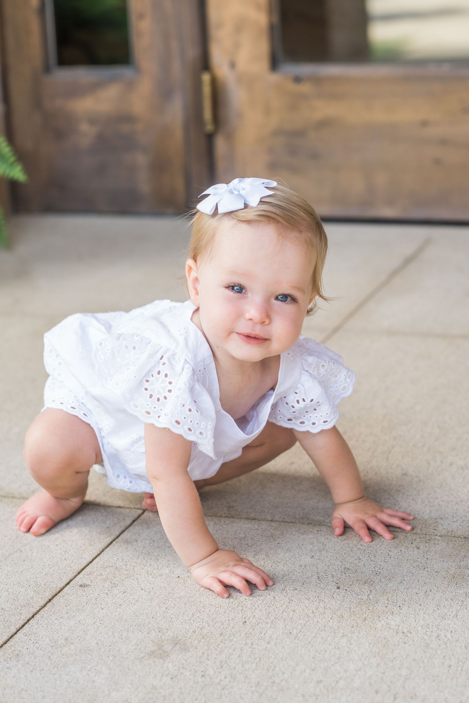 Adorable baby girl in white eyelet romper and bow headband crawling on outdoor patio pavers.
