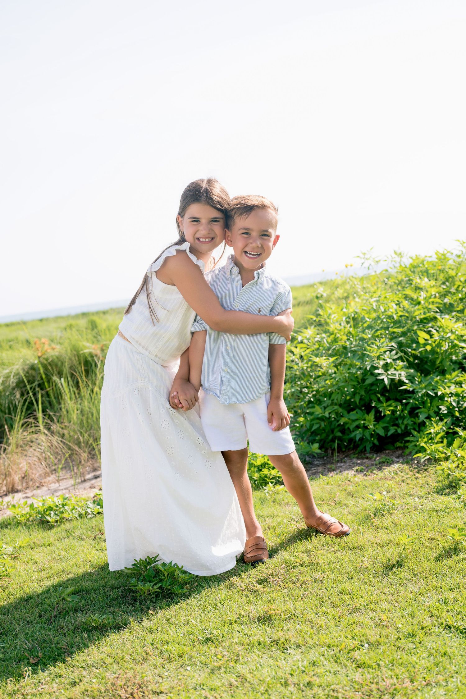 Two happy children hugging outdoors on green grass, girl in white dress embracing smiling boy in light blue shirt.