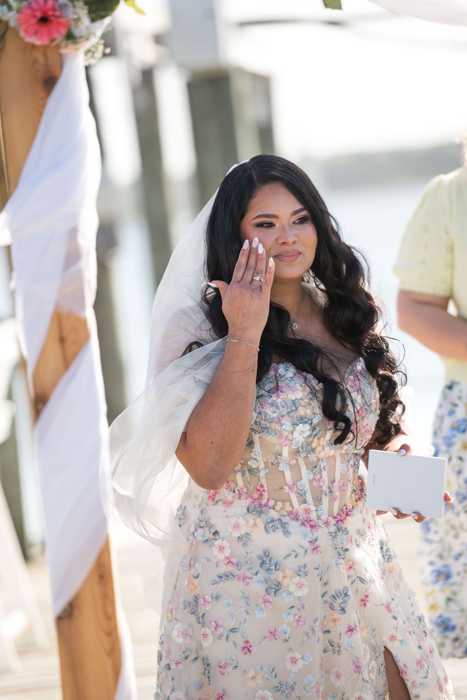 Emotional bride in floral gown and veil wiping tears at outdoor waterfront wedding ceremony.