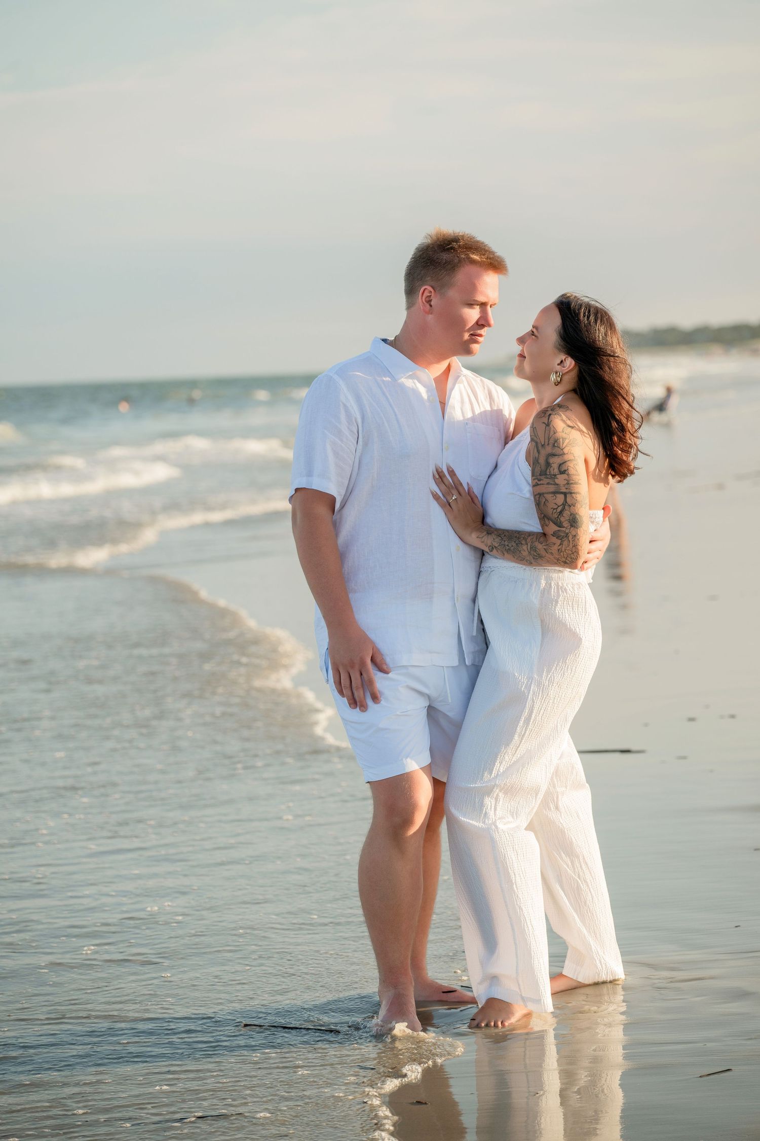Couple in white outfits embracing on a sandy beach with ocean waves, sharing a romantic moment at sunset.