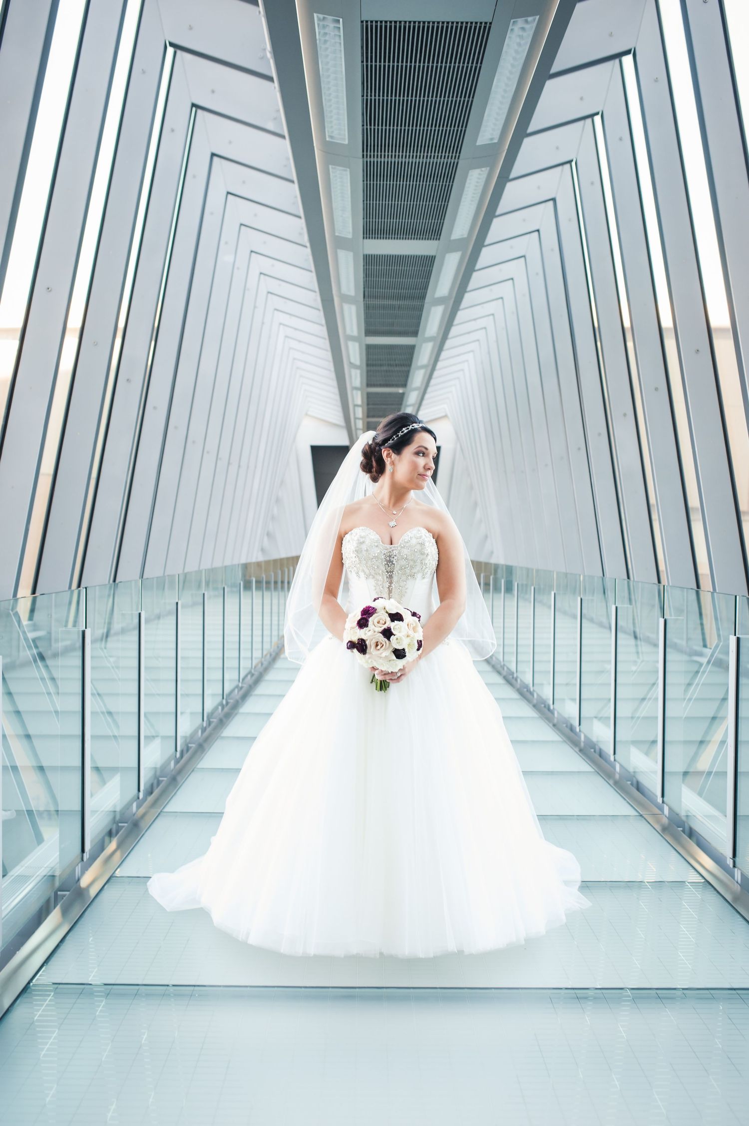 A bride in a white ballgown poses in a modern glass-walled corridor with geometric architectural details.