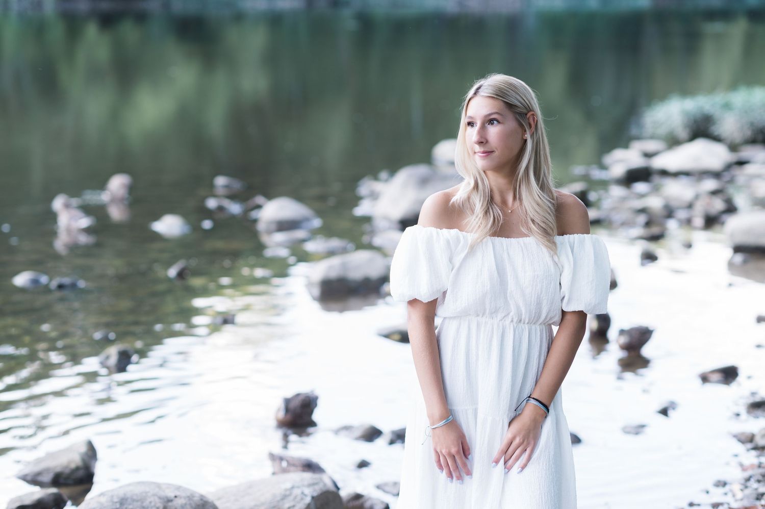 A couple in white outfits poses together by a scenic lake with rocks and trees in the background.