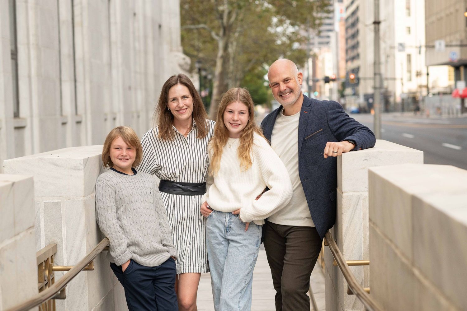 A smiling family of four poses outdoors on a city street, wearing casual stylish outfits in a bright urban setting.