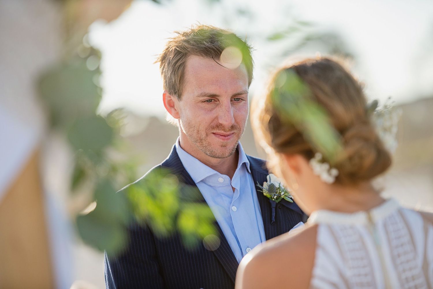 Groom in navy suit gazing lovingly at bride during outdoor wedding ceremony with greenery in background.