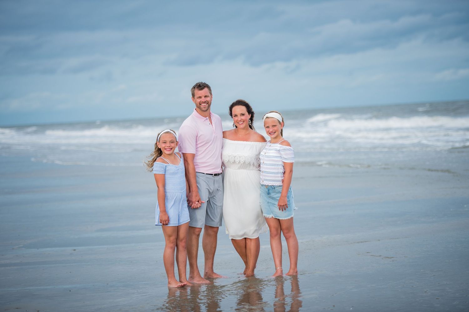 A family wearing white outfits poses together on a sunny beach with ocean waves in the background.