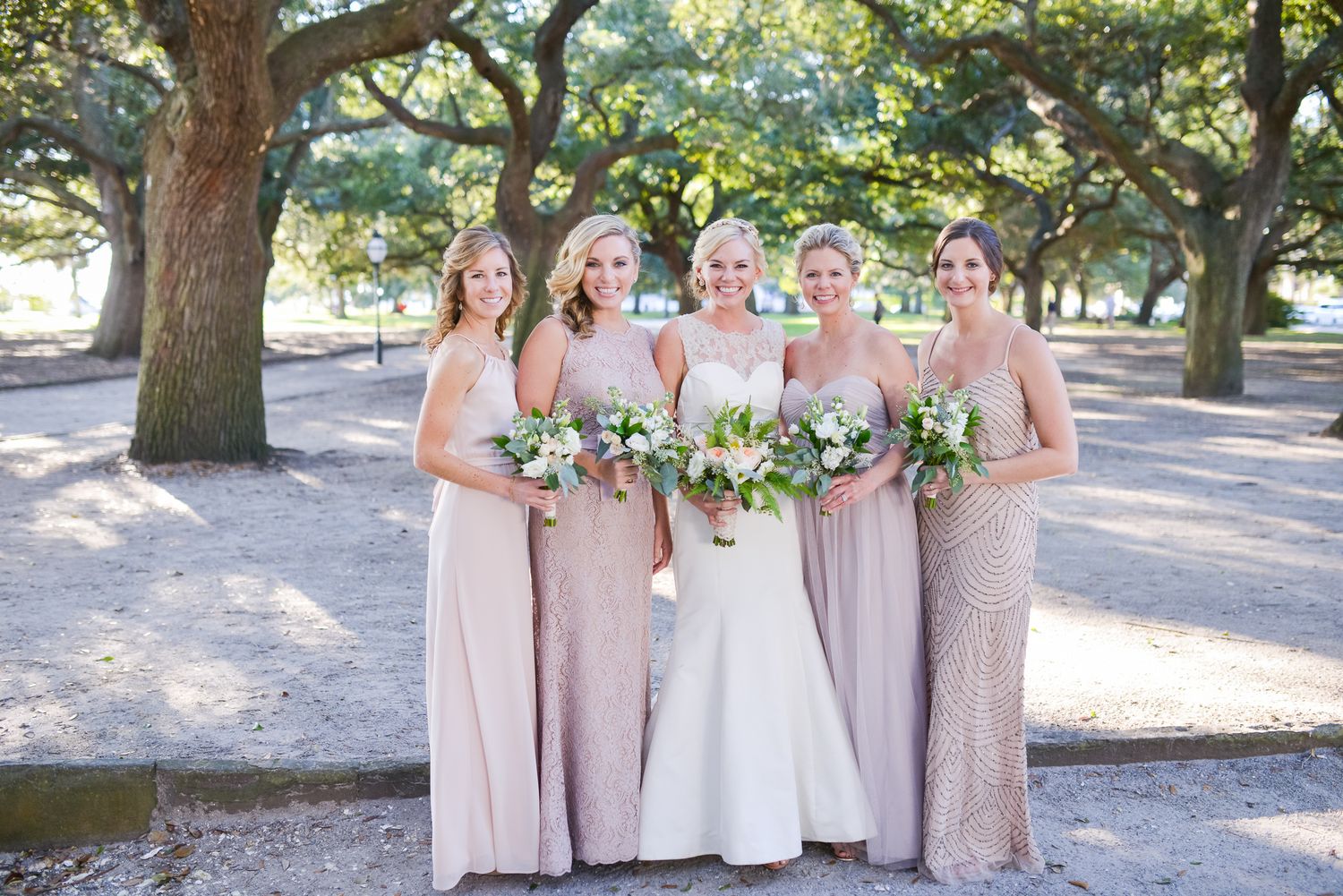 Wedding party in blush pink dresses poses under oak trees in an outdoor garden setting.