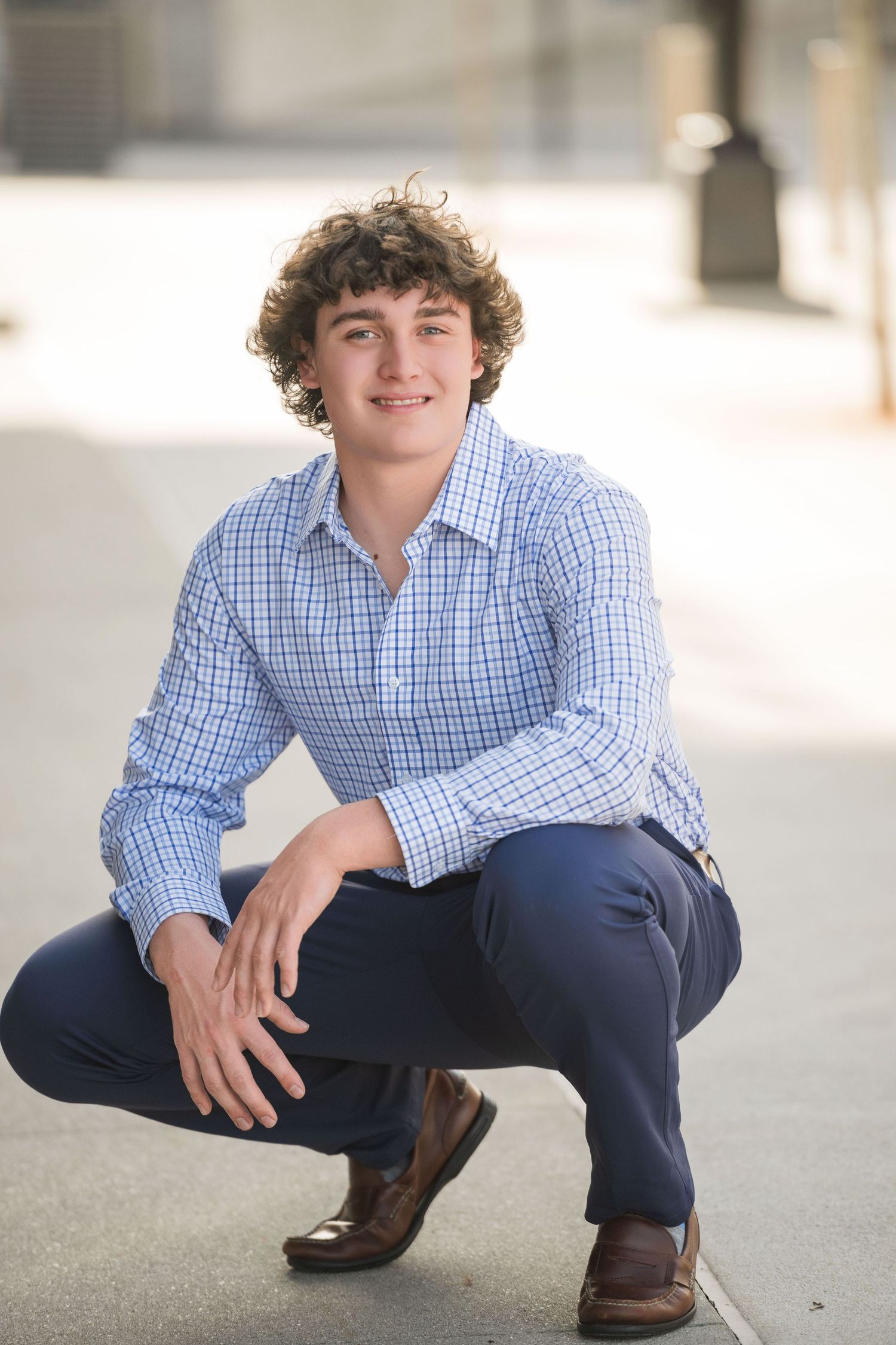 A person in a blue checkered shirt and navy pants poses casually while crouching on a concrete surface.