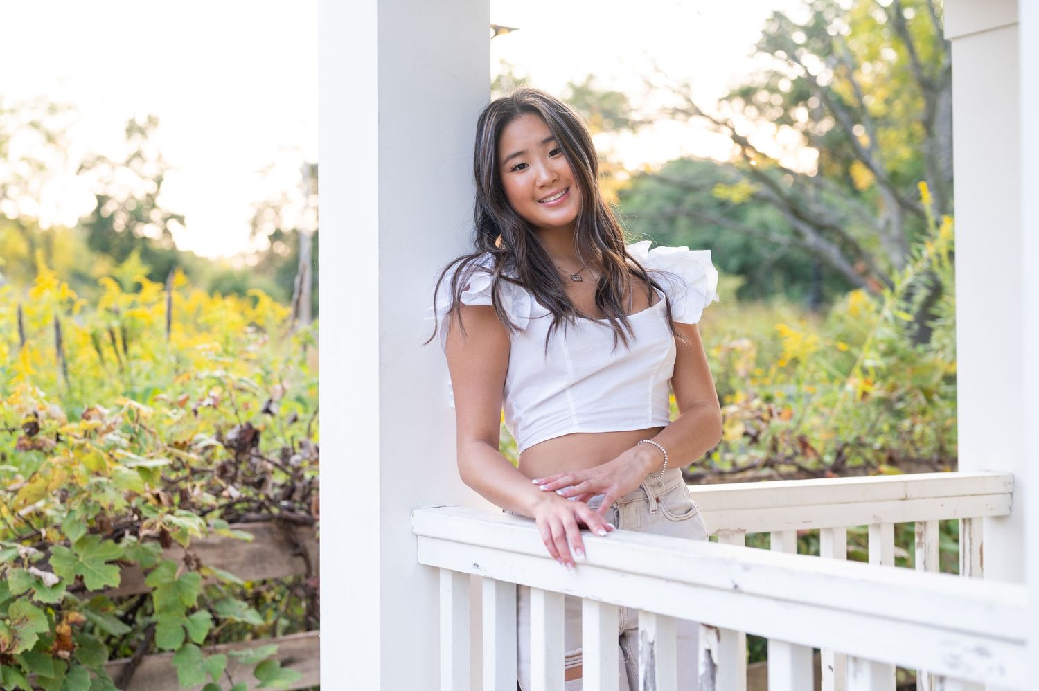A person in a white top leans against a white porch railing surrounded by lush garden foliage at sunset.