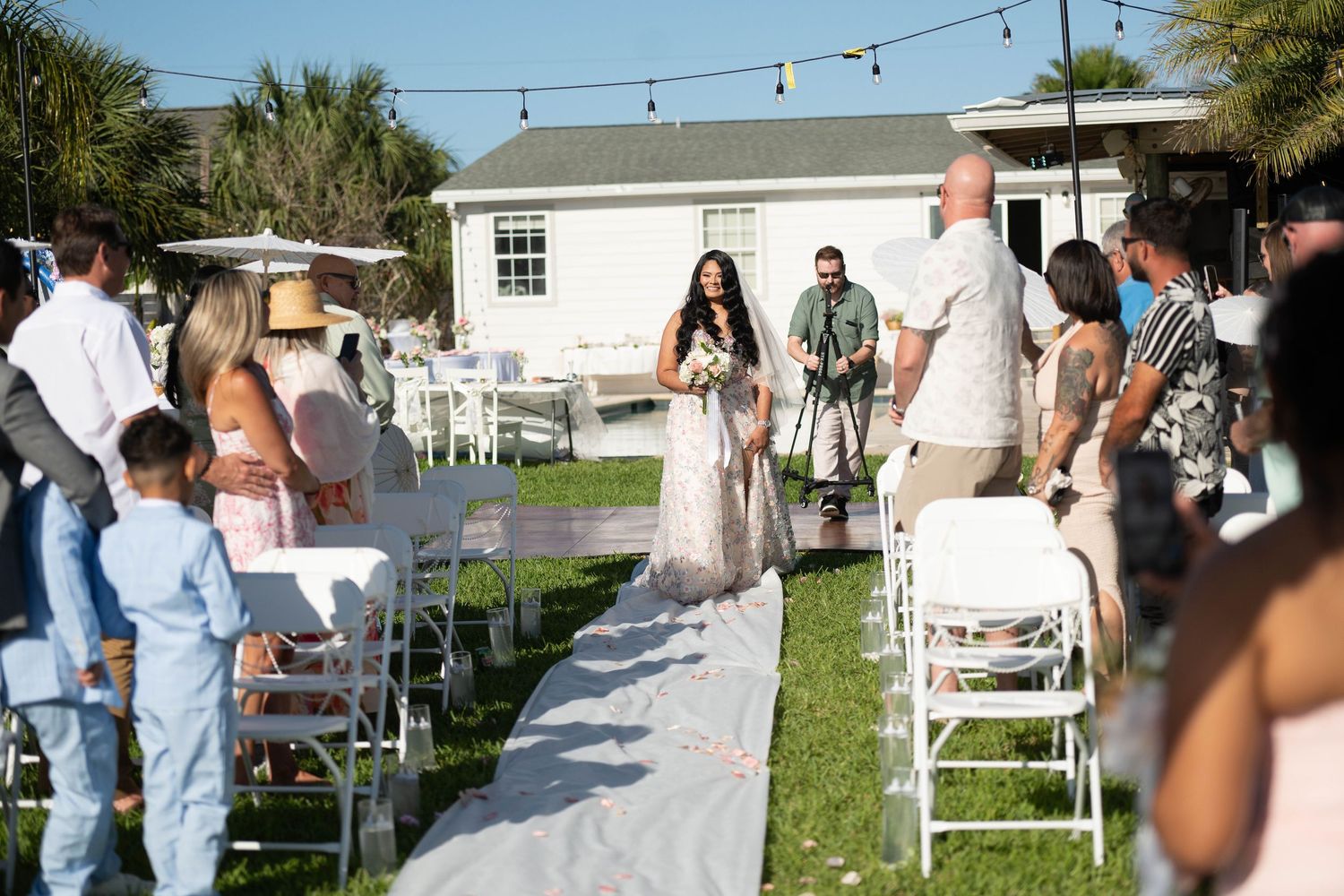 Bride walks down outdoor aisle with escorts at a sunny beach wedding ceremony with white chairs and string lights.