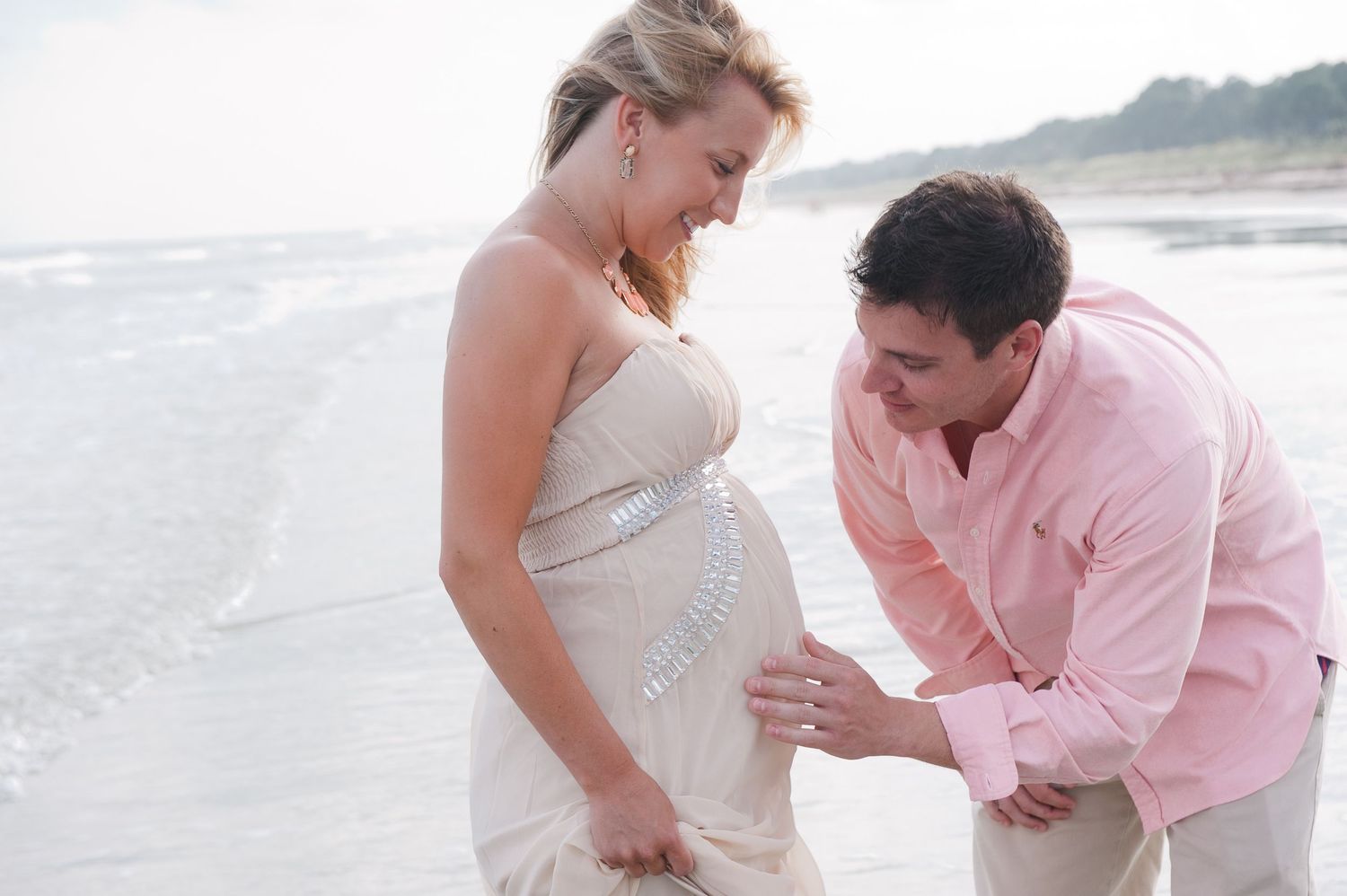 Pregnant woman in white dress with husband kneeling at beach, touching baby bump during maternity photoshoot.