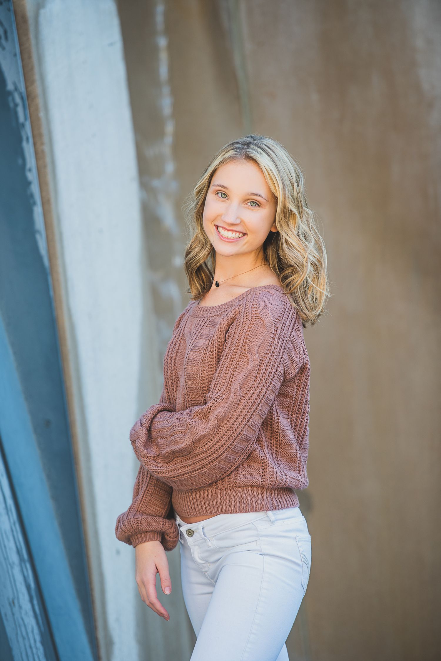 Smiling blonde woman in a pink knit sweater and white jeans posing against a textured wall.