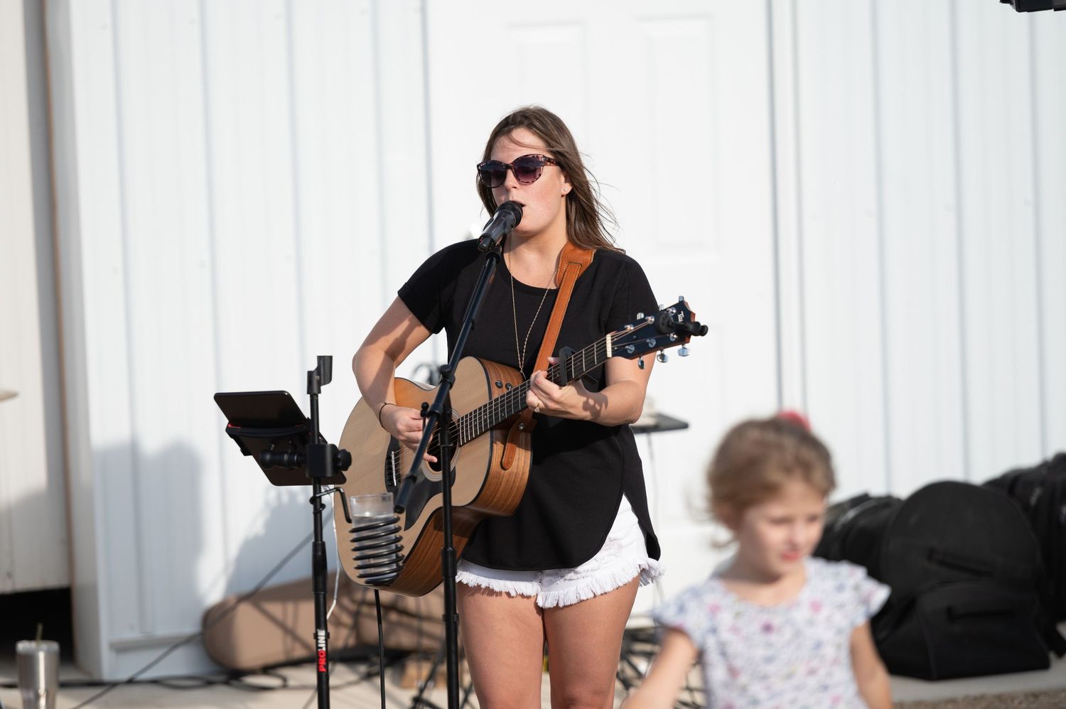 Musicians perform on acoustic guitars at an outdoor venue with white walls and a casual summer atmosphere.