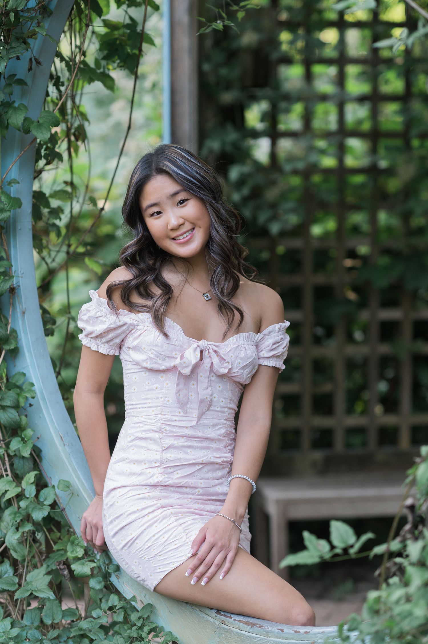 A person in a white ruffled dress poses gracefully in a garden setting with wooden lattice and greenery in the background.
