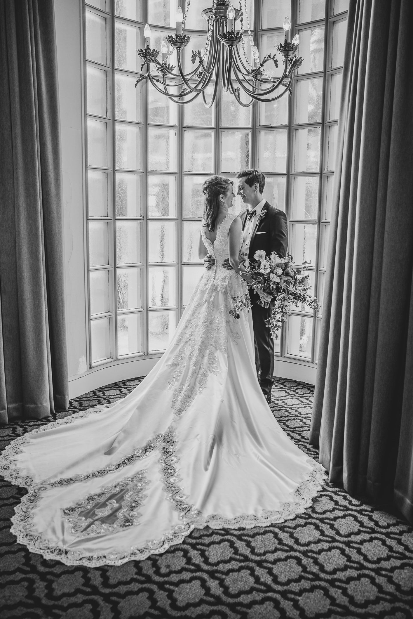 A bride and groom stand together in an elegant room with large windows and a cathedral-length lace wedding dress train. Athletic Club.