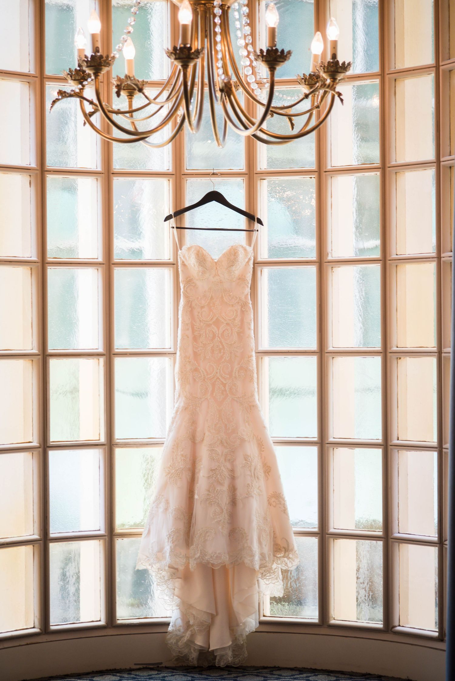 A blush lace wedding dress hangs in a bay window beneath an elegant bronze chandelier at the Athletic Club, Columbus, Ohio