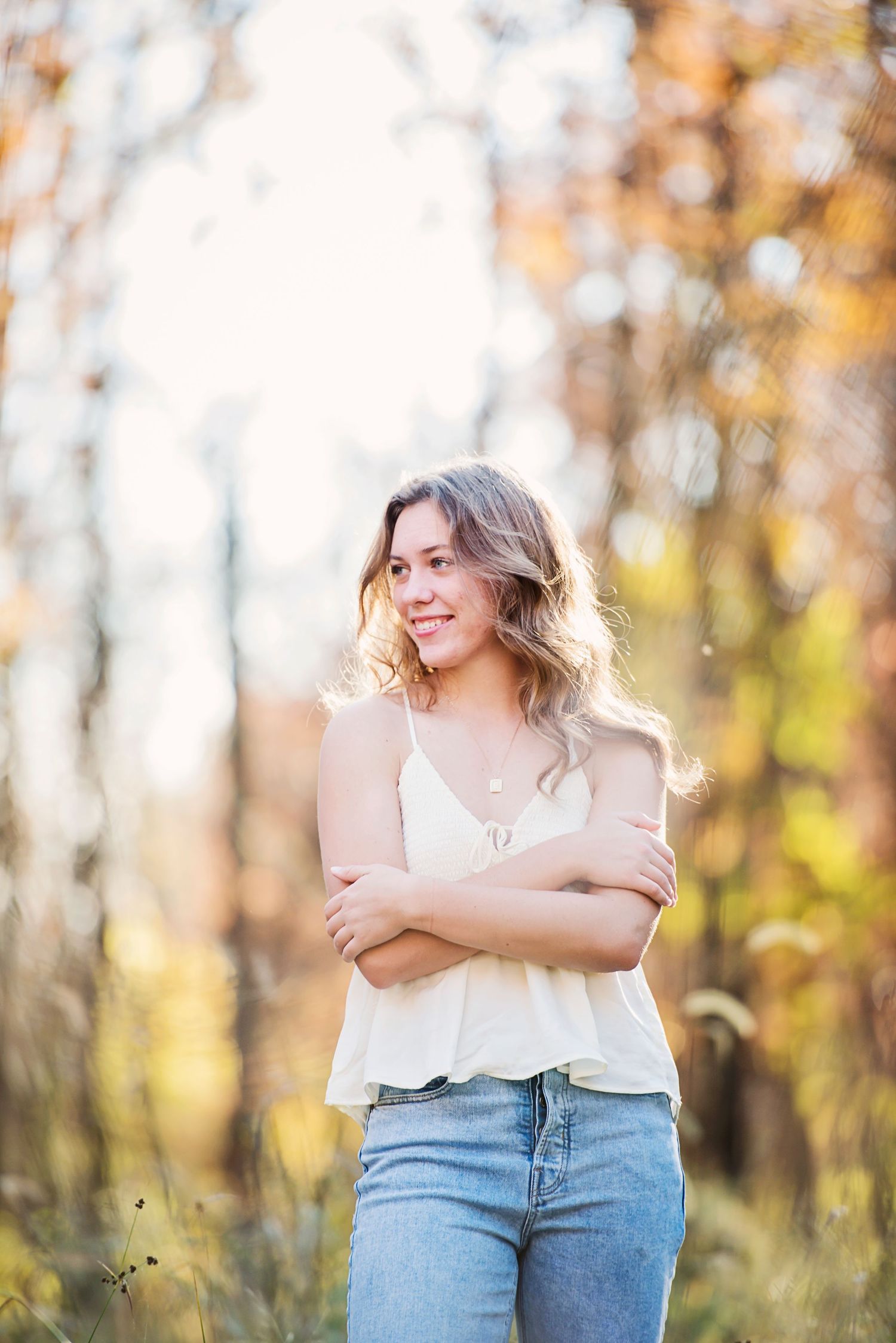 Senior Pictures white camisole and jeans smiling with arms crossed in a sunlit autumn forest setting.