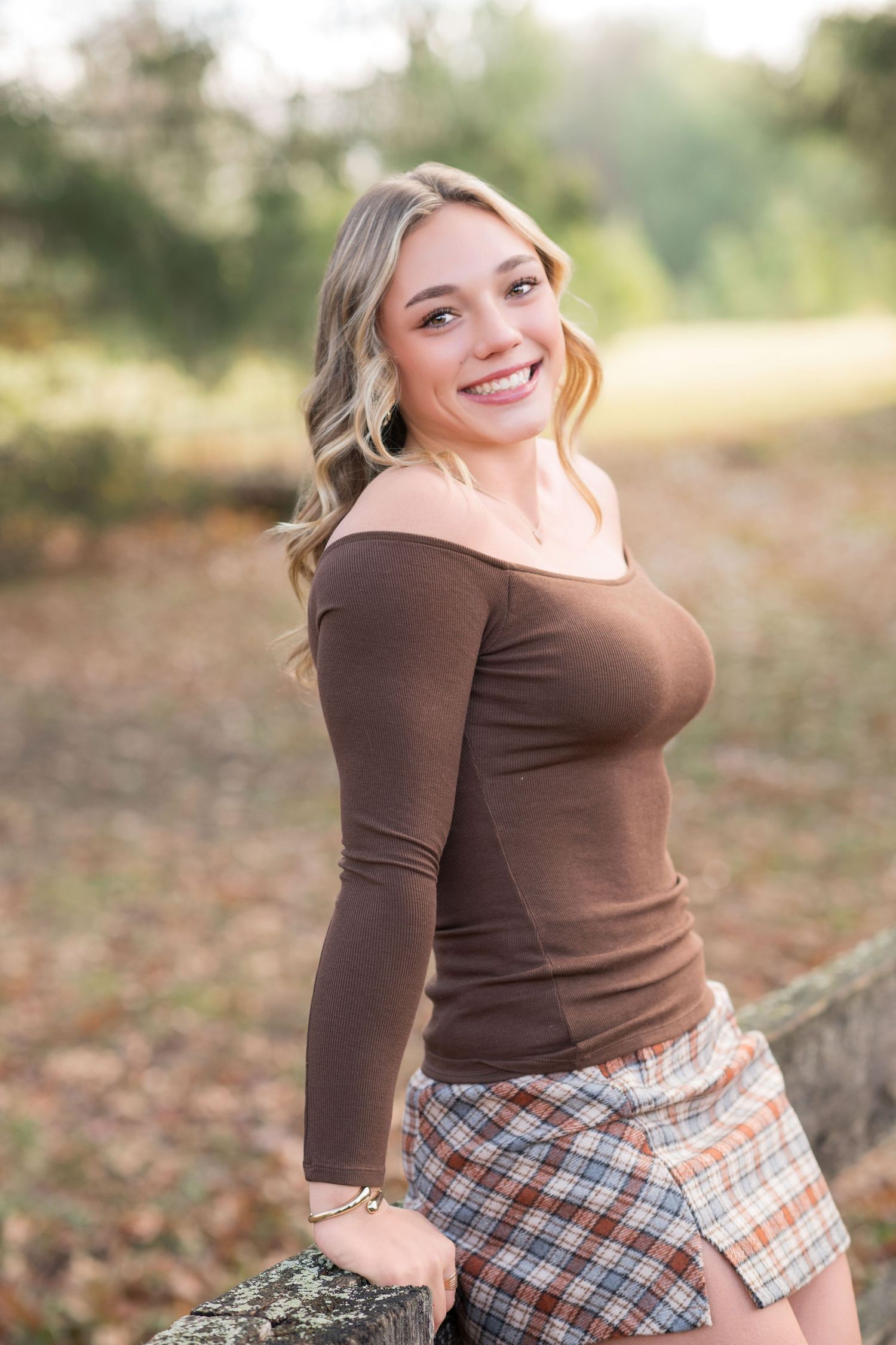 Person in brown long sleeve top and plaid skirt poses outdoors during autumn season.