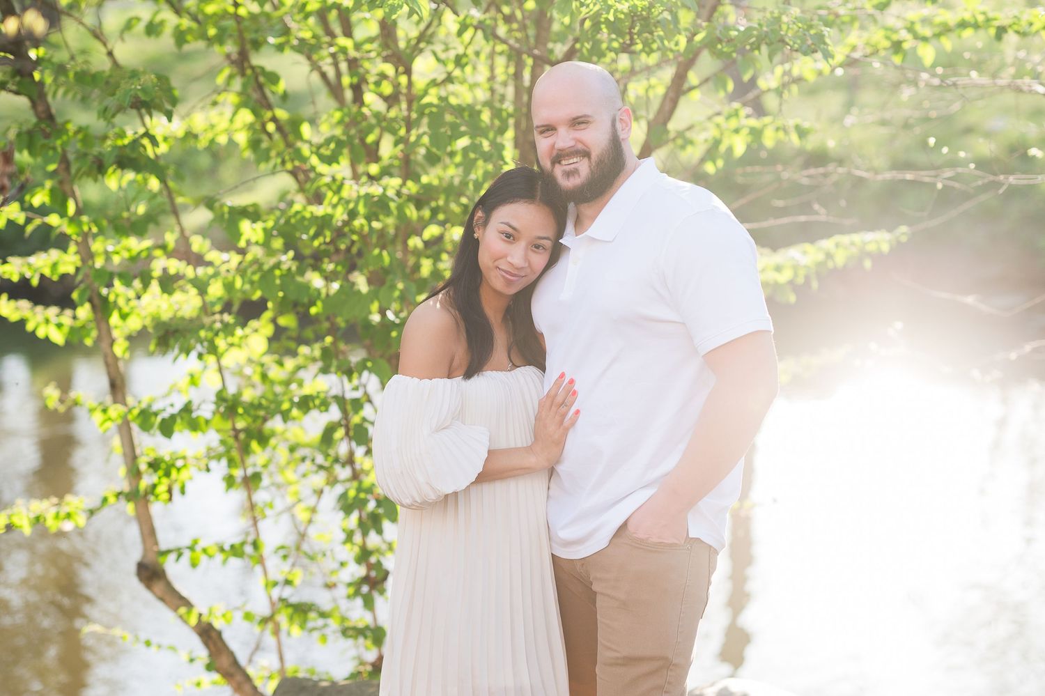 Romantic engagement photos in sunlit garden with couple in white and neutral outfits.