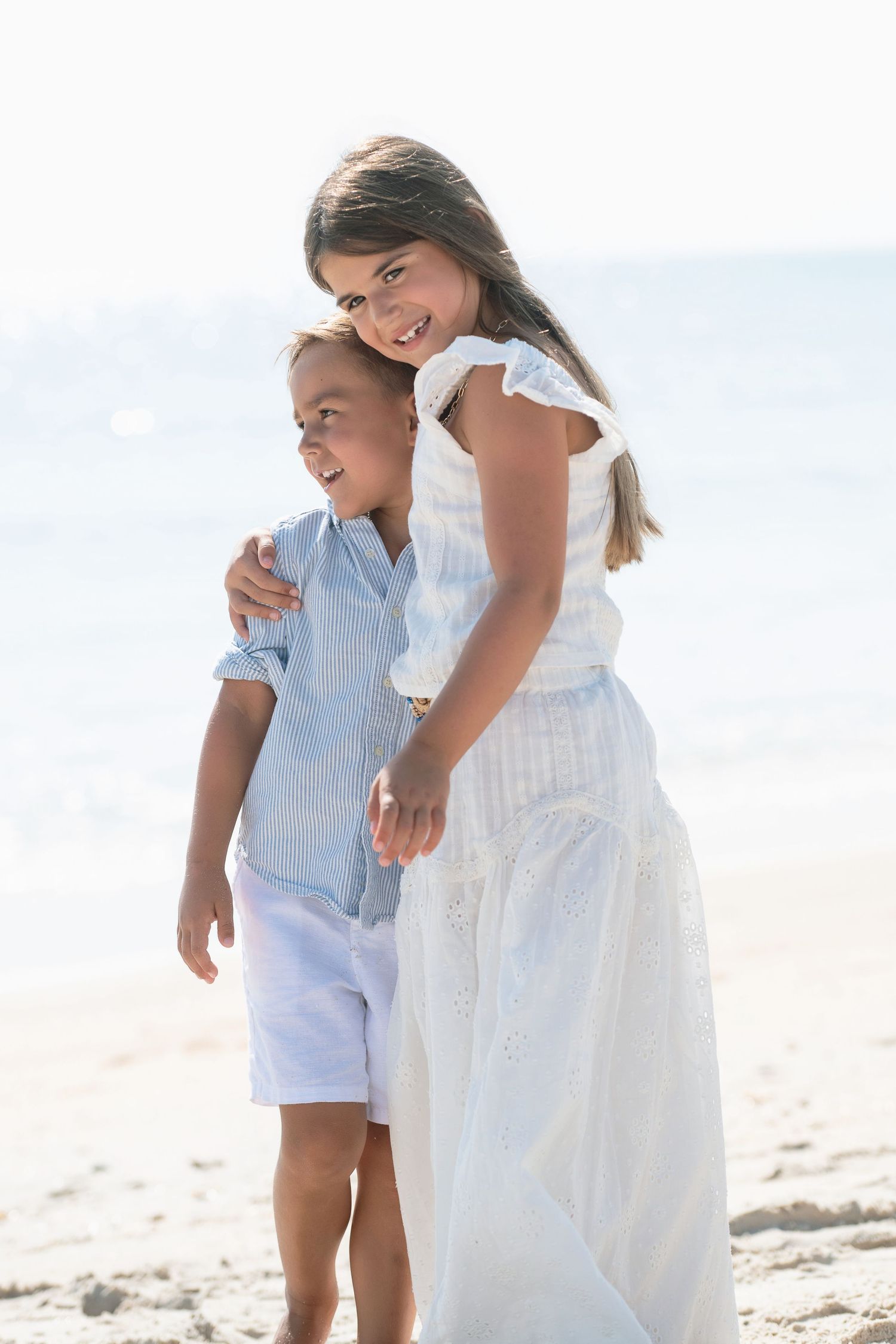 Two children smile and embrace on a sunny beach, girl in white dress hugging younger boy in blue shirt and white shorts.