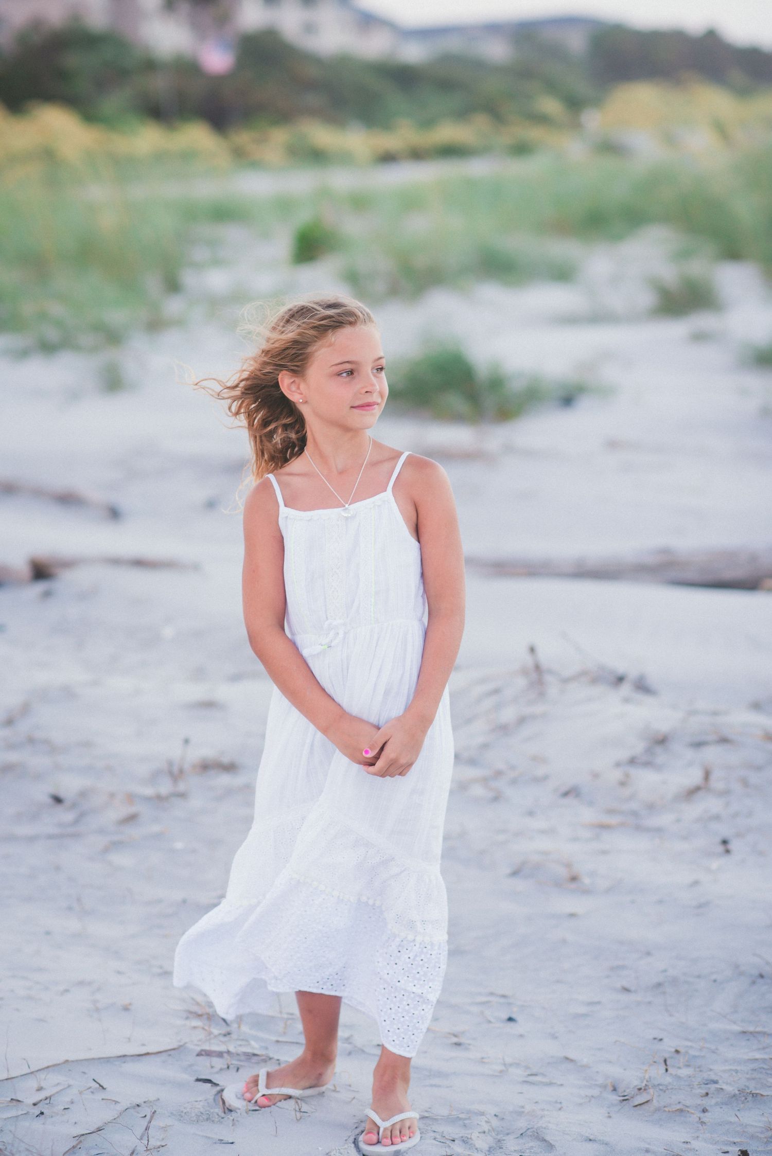 Young girl in  Hilton Head white sundress standing on sandy beach with wind blowing her hair. Family photo session.