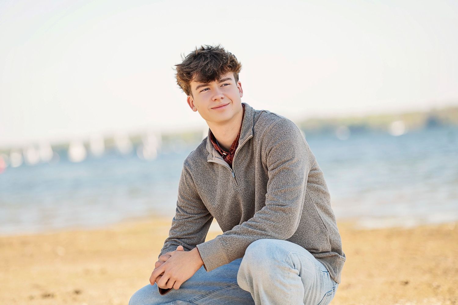 A young man in a gray hoodie and jeans sits on a sandy beach, smiling warmly with the ocean in the background.