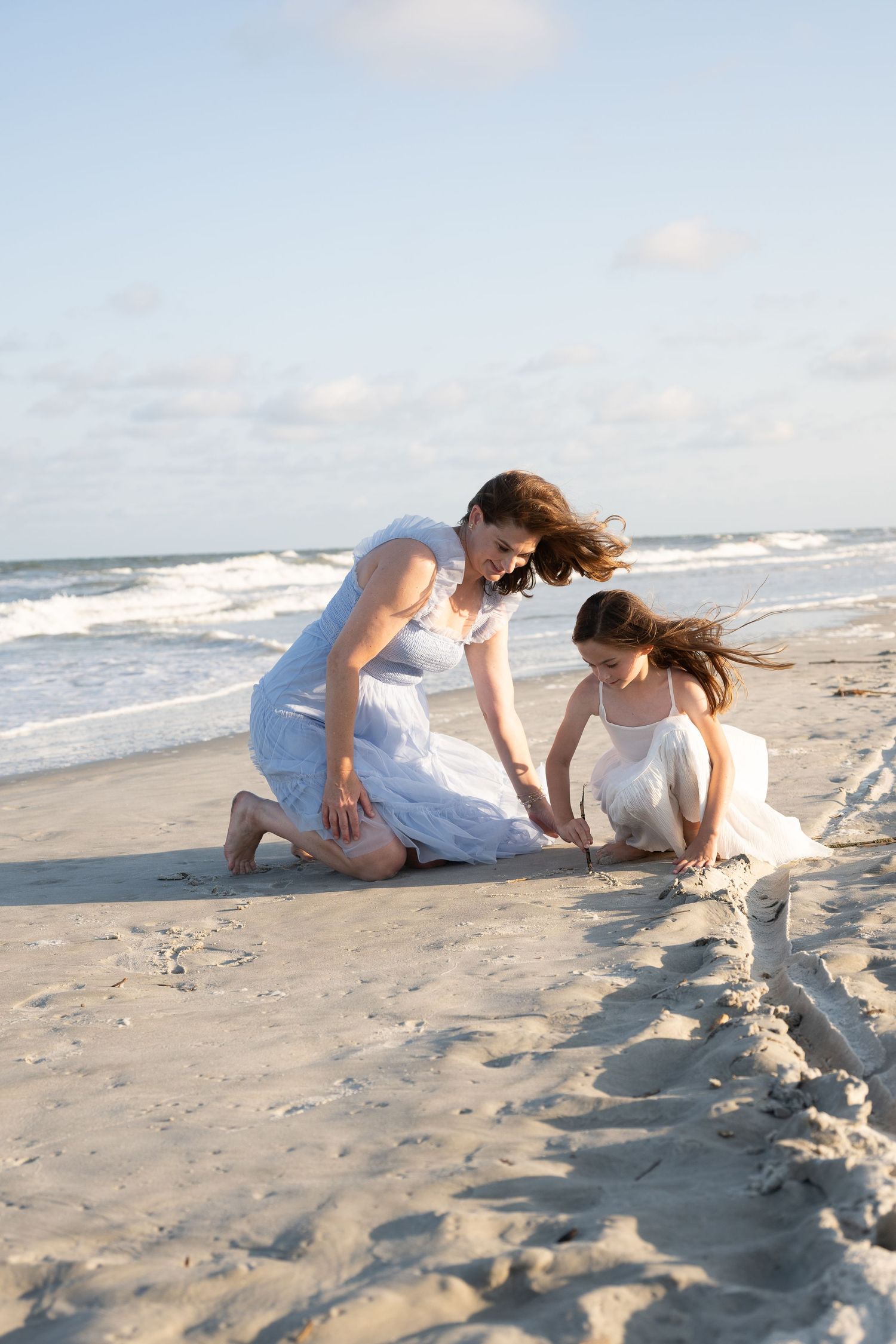 Mother and daughter in white dresses playing in the sand on a beach with ocean waves in the background.