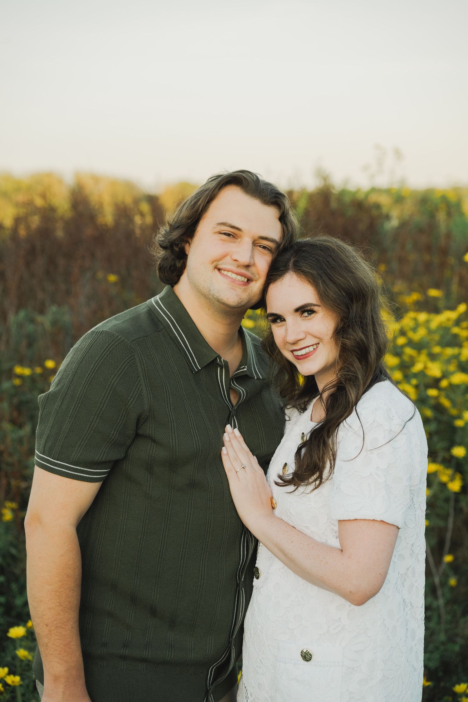 A smiling couple poses together in a sunflower field at golden hour, woman in white dress, man in green polo shirt.