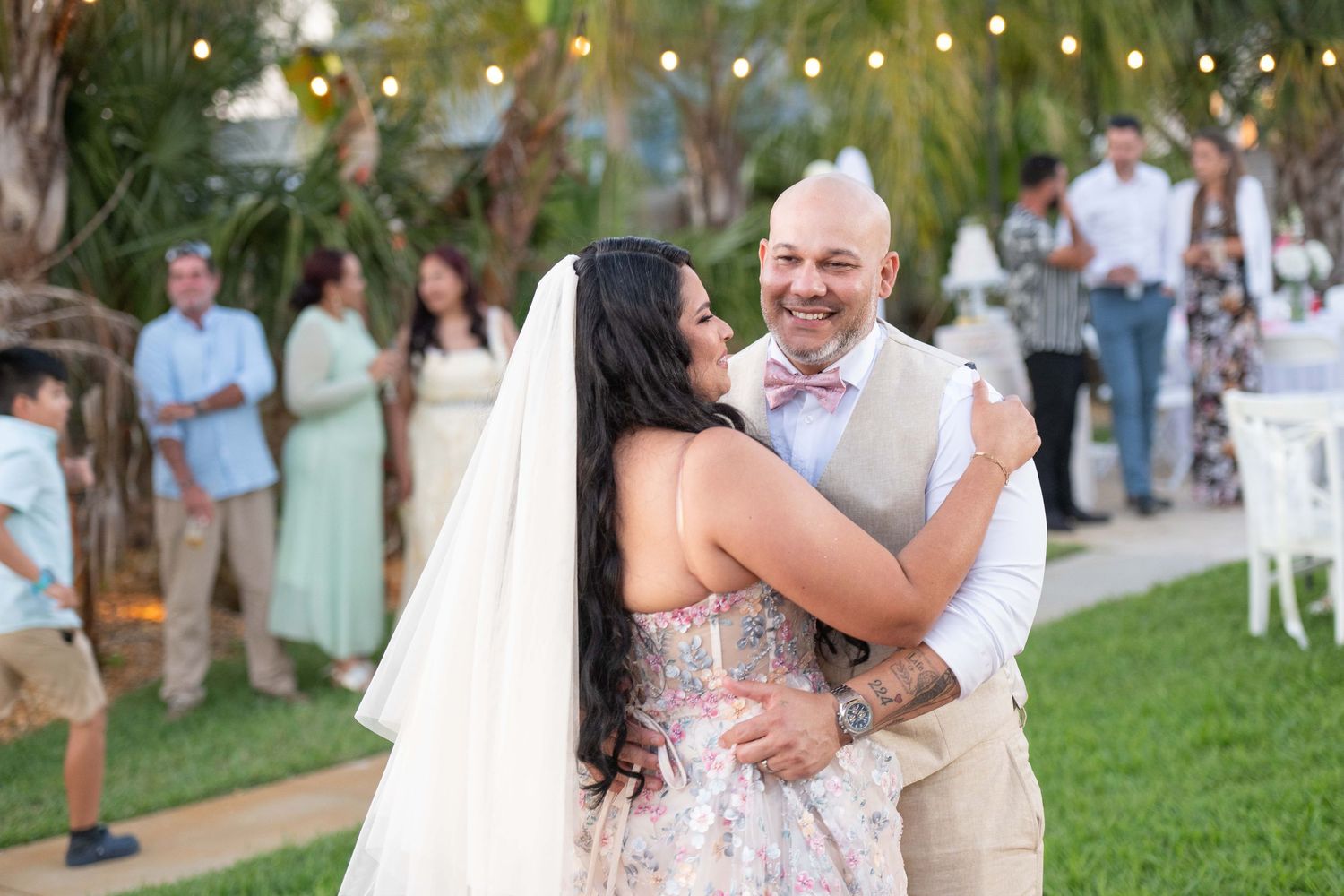 Bride and groom share first dance at outdoor wedding reception with string lights and guests in background.