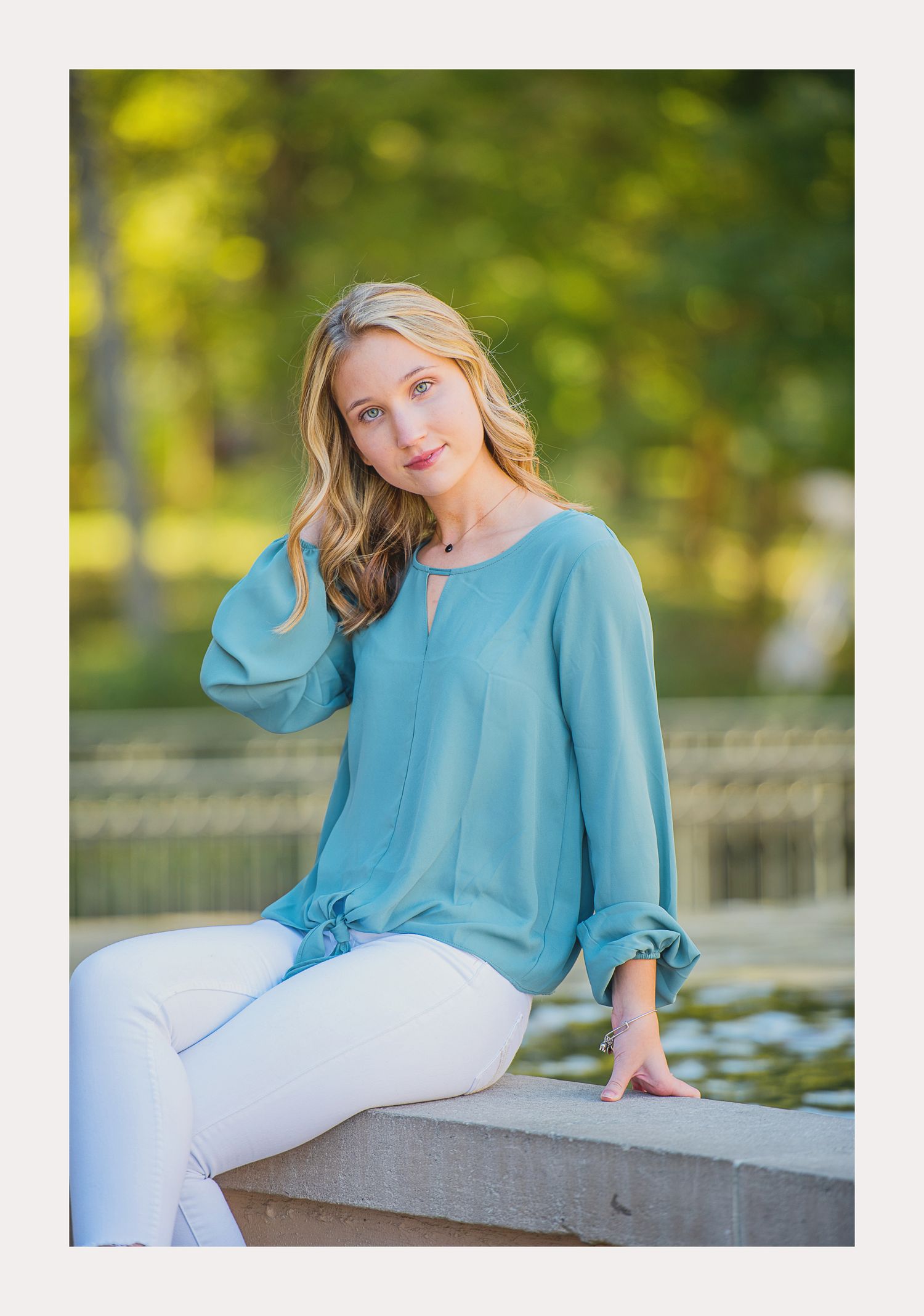 Young blonde woman in teal blouse and white jeans sits outdoors near a fountain with lush green bokeh background.