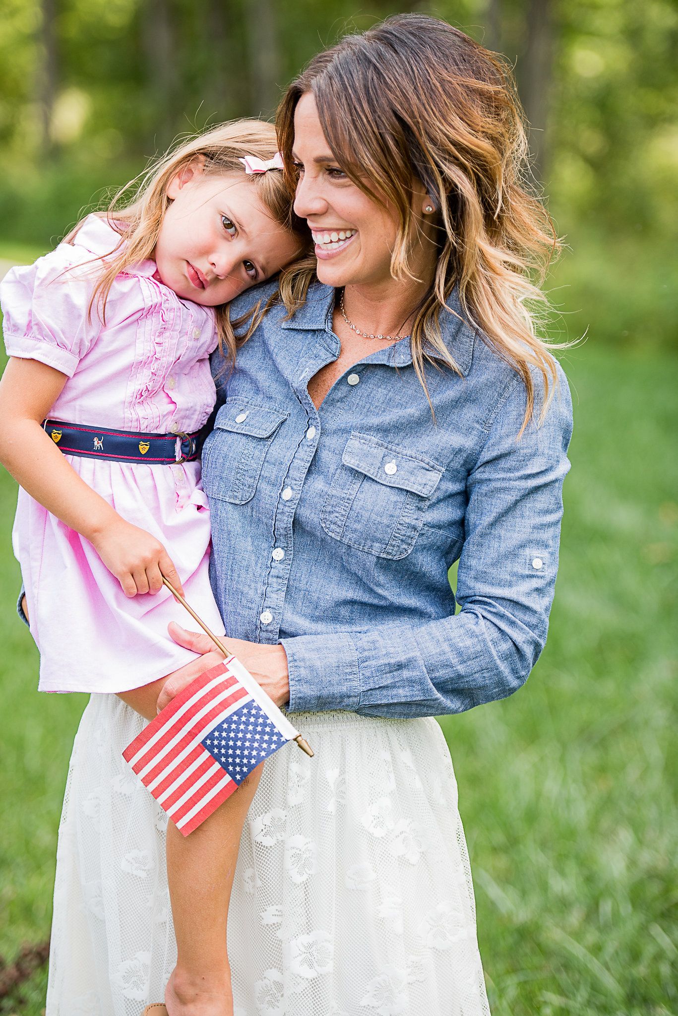 A smiling woman in a chambray shirt holds a little girl in a pink dress clutching a small American flag outdoors.