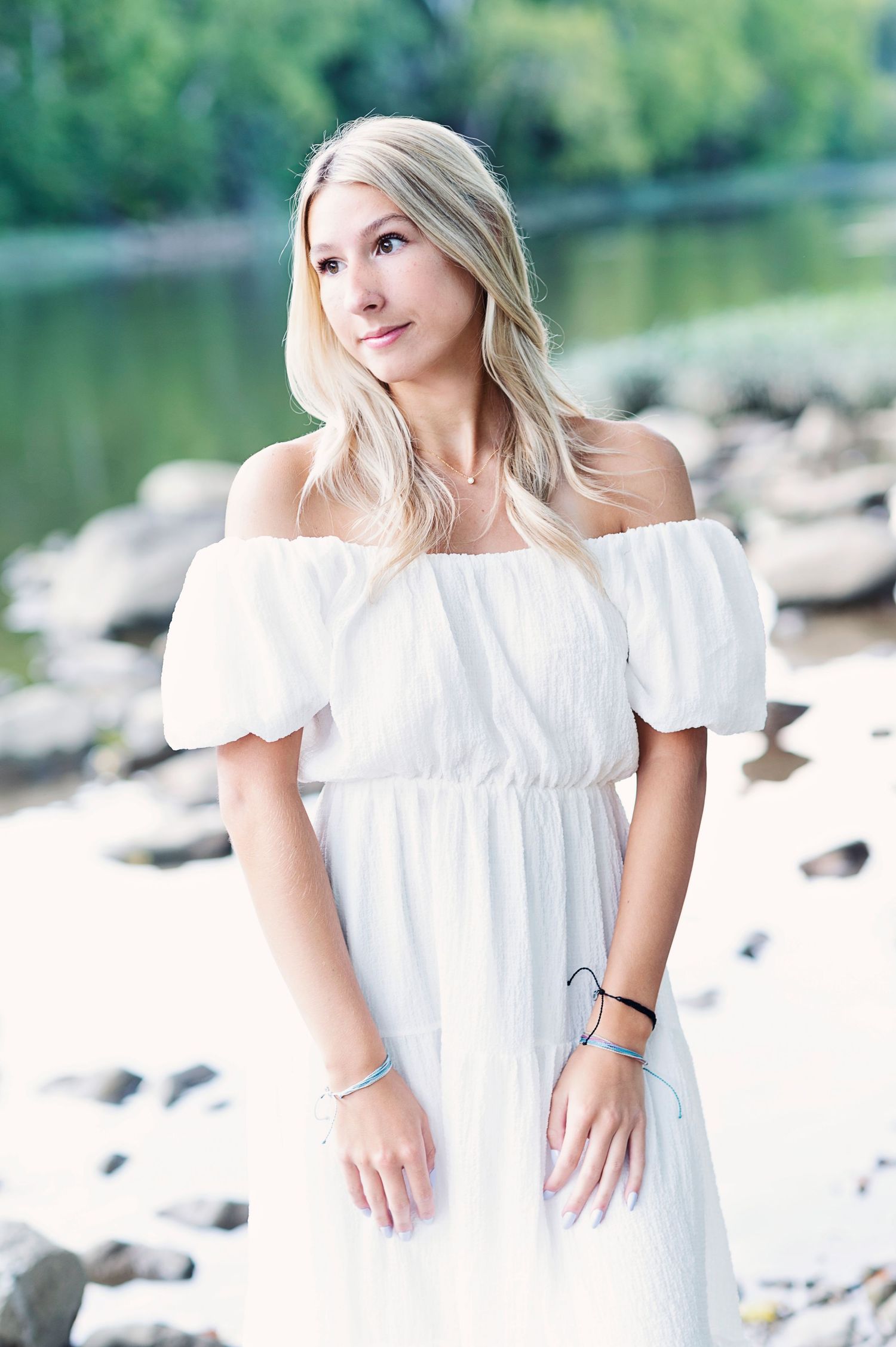 Waterfront portrait in white off-shoulder dress with rocks and lake in background.