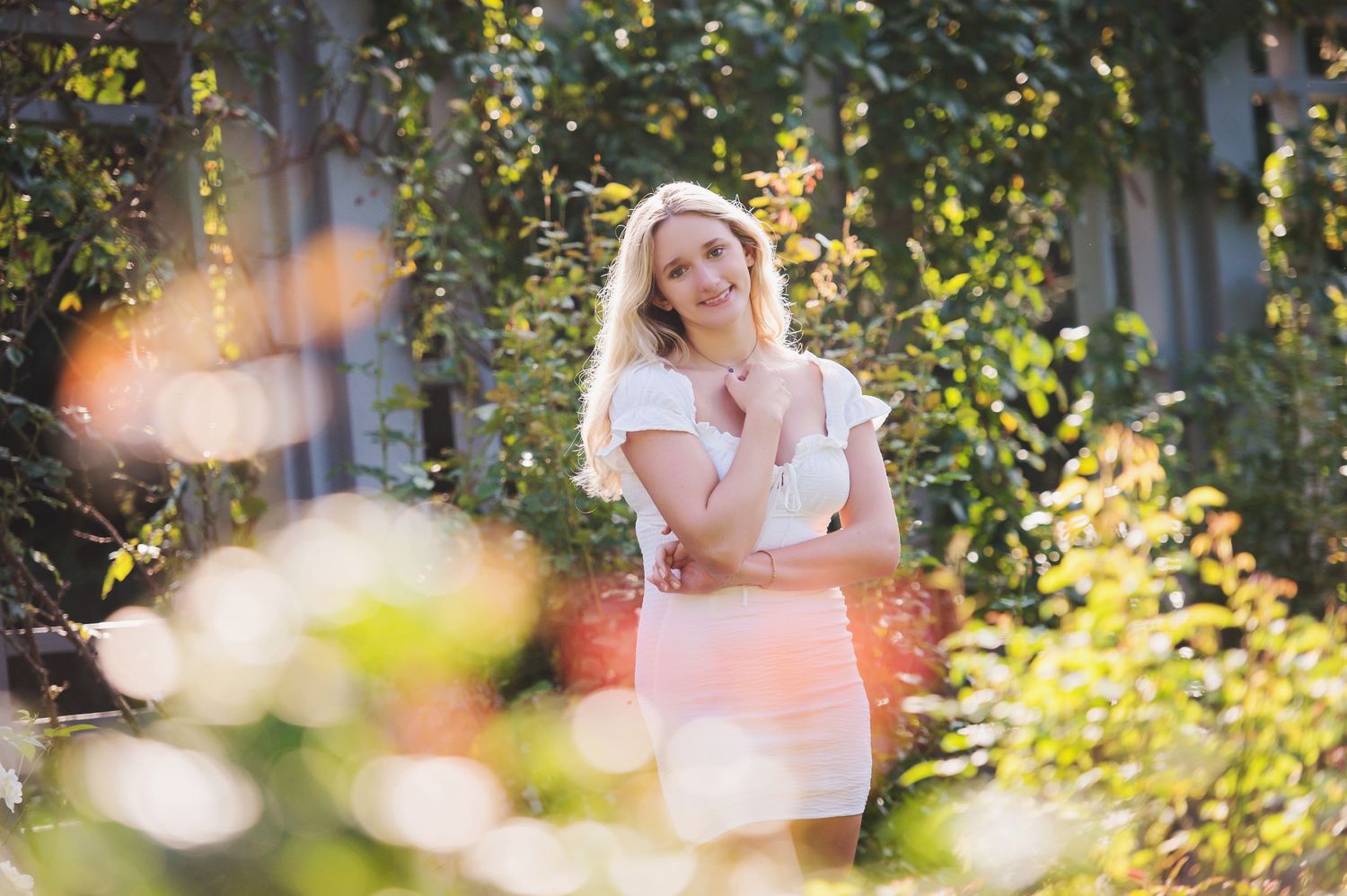A person in a white dress poses in a sunlit garden setting with lush greenery and bokeh effects in the background.