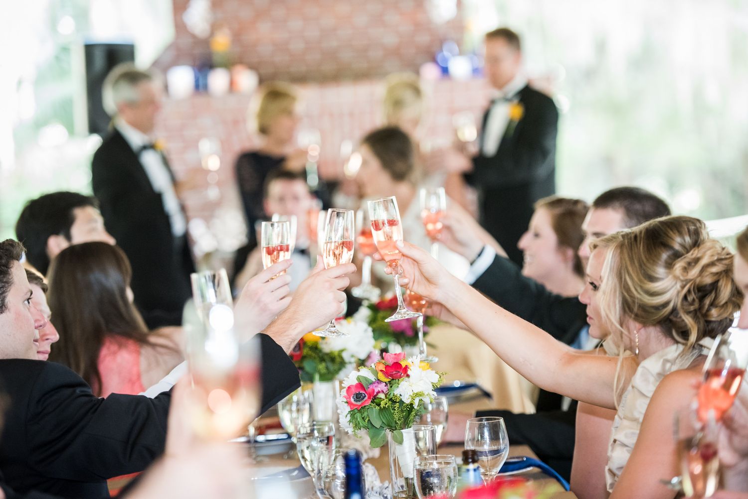 Wedding guests raise champagne glasses in celebration during an elegant indoor reception with floral centerpieces.