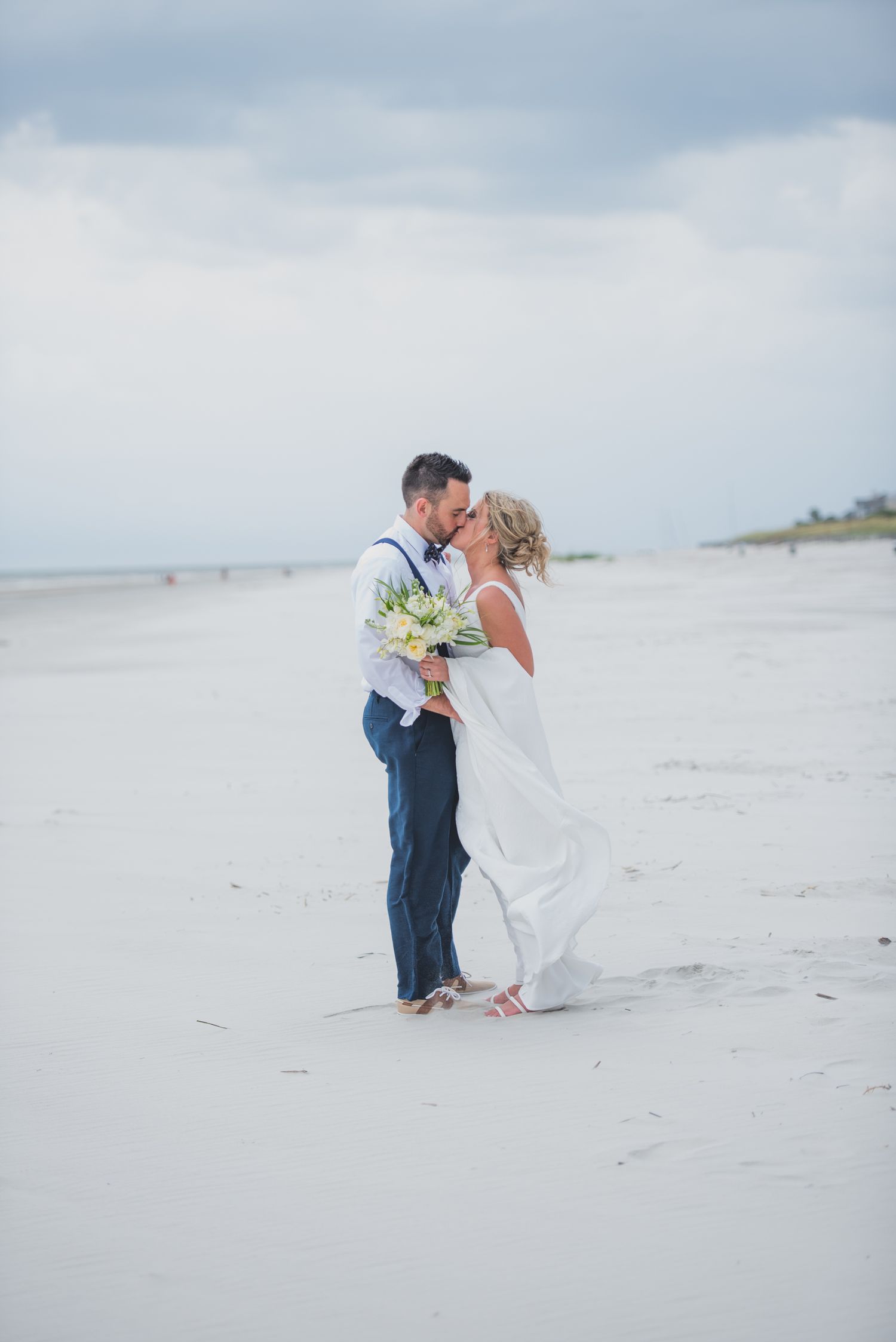 A romantic beach wedding scene with a couple embracing on white sand as waves roll in the background.