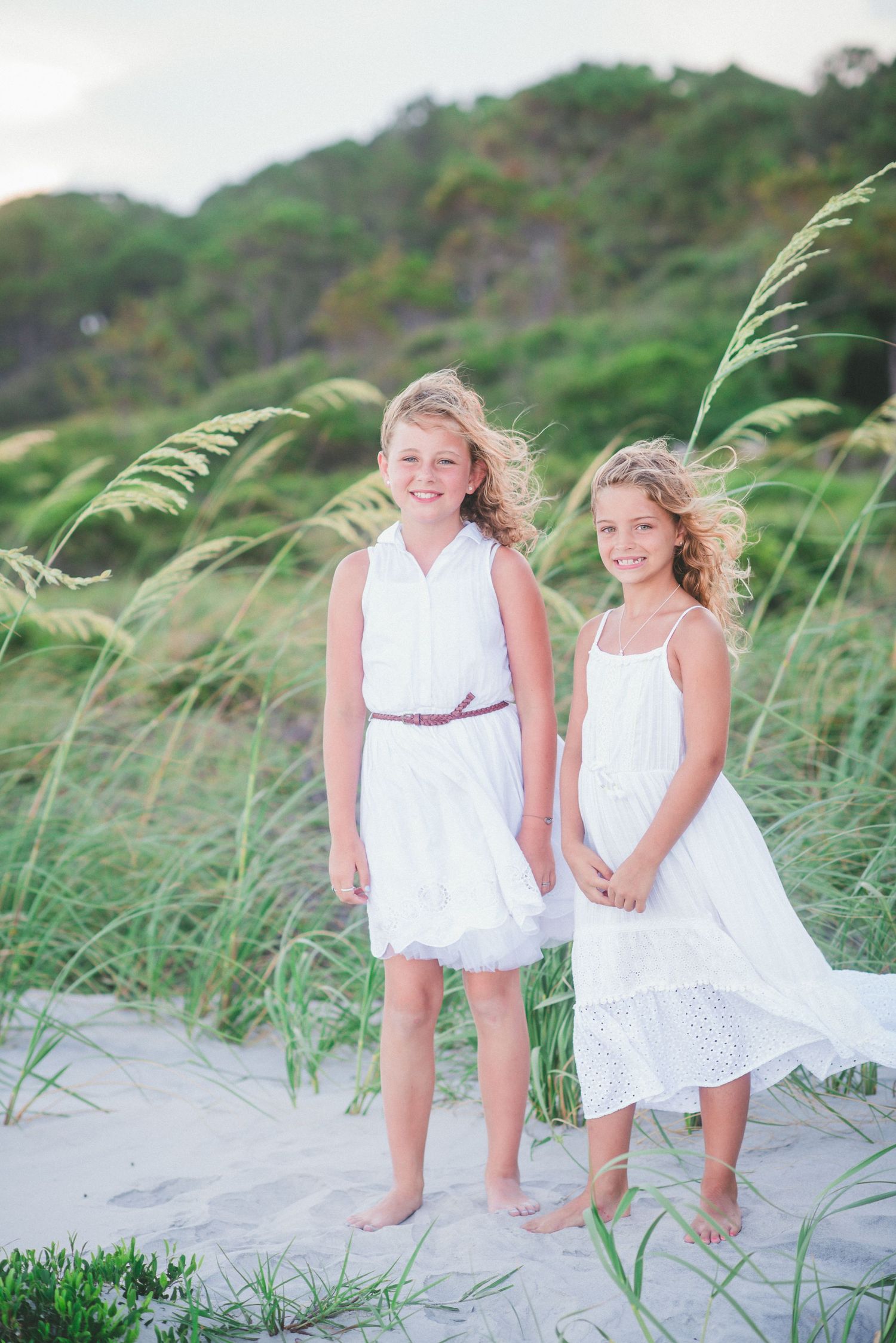 Two children in white dresses holding hands while walking through beach grass at sunset. St. Augustine, Palm Coast, Florida photo session.