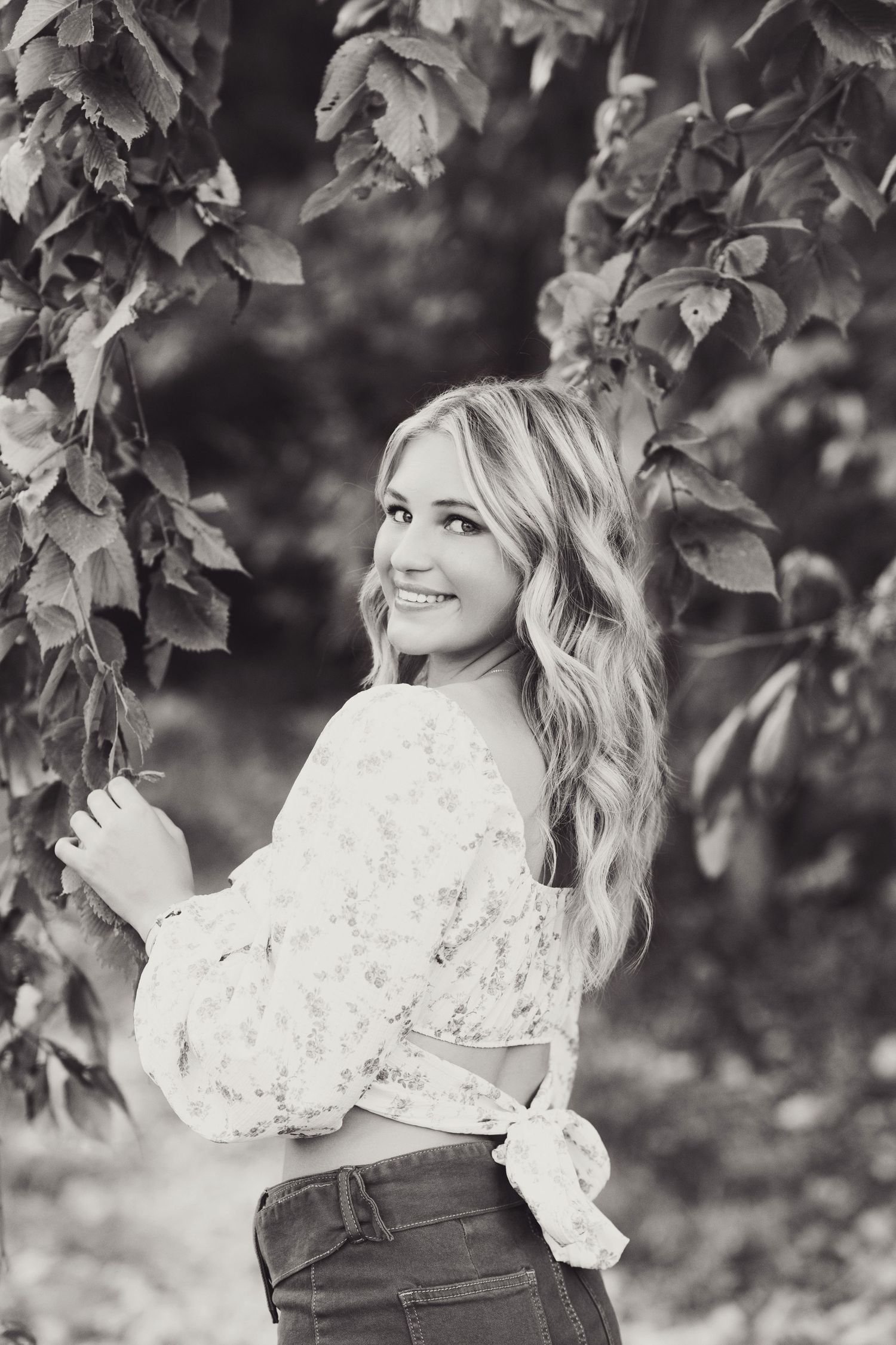 Black and white portrait series showing a woman in a white lace top and jeans posing among tree leaves outdoors.