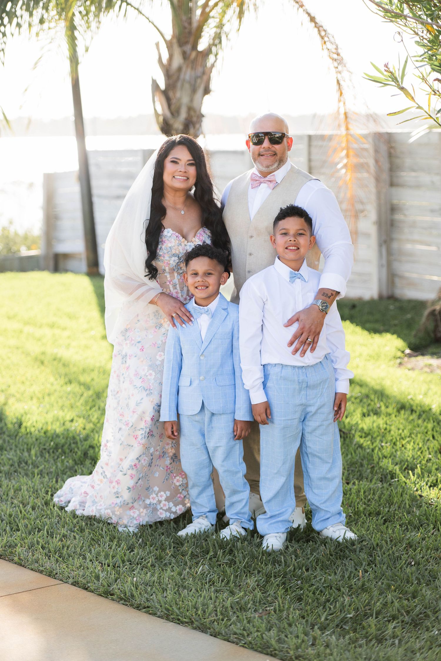 A wedding family portrait with bride in floral gown, groom in tan suit, and two boys in light blue suits on green lawn.