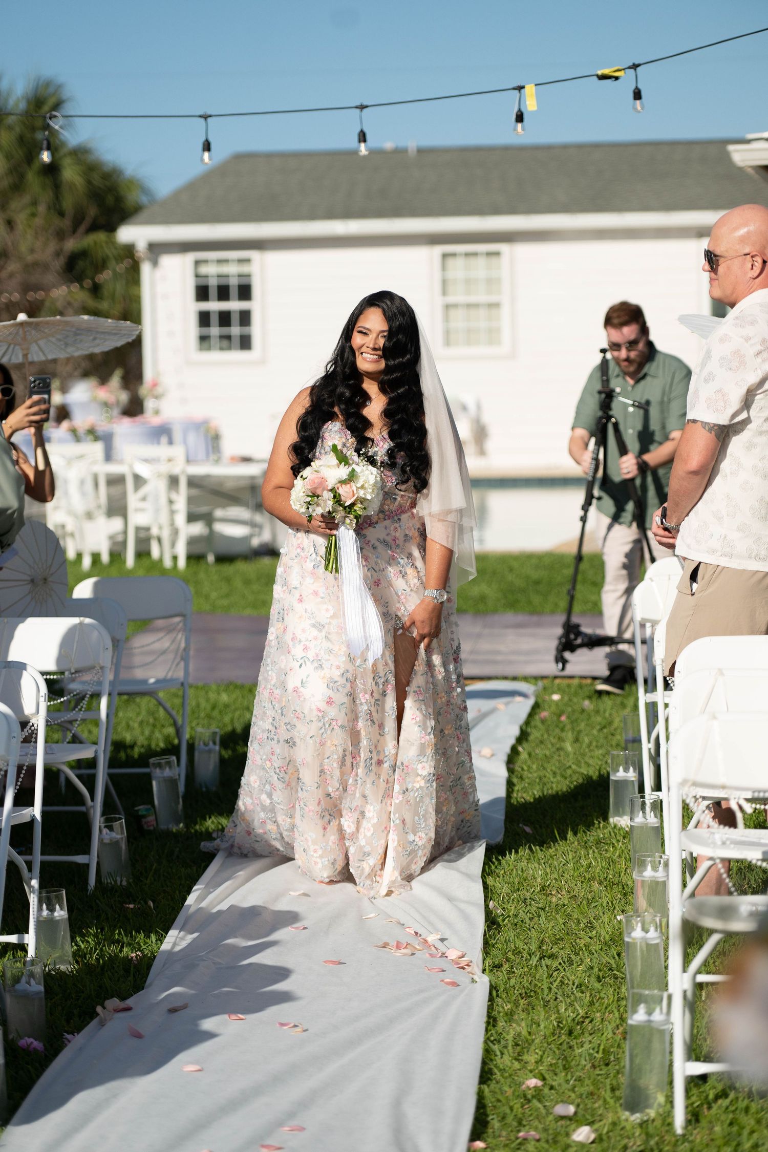 Bride walks down outdoor aisle in floral gown with veil, holding bouquet at backyard wedding ceremony.