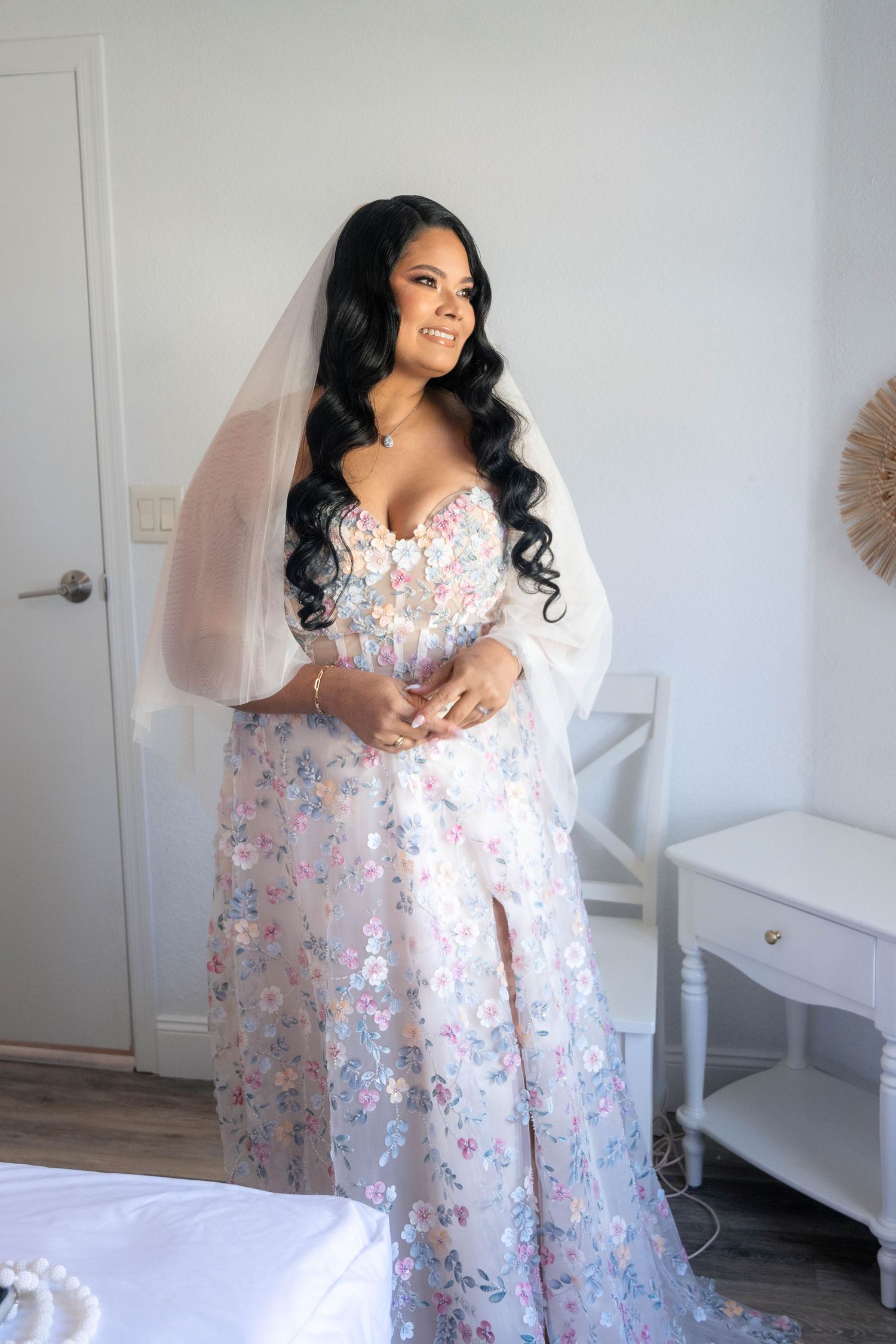 Bride in floral wedding gown with veil and sheer sleeves smiling in bright white room.
