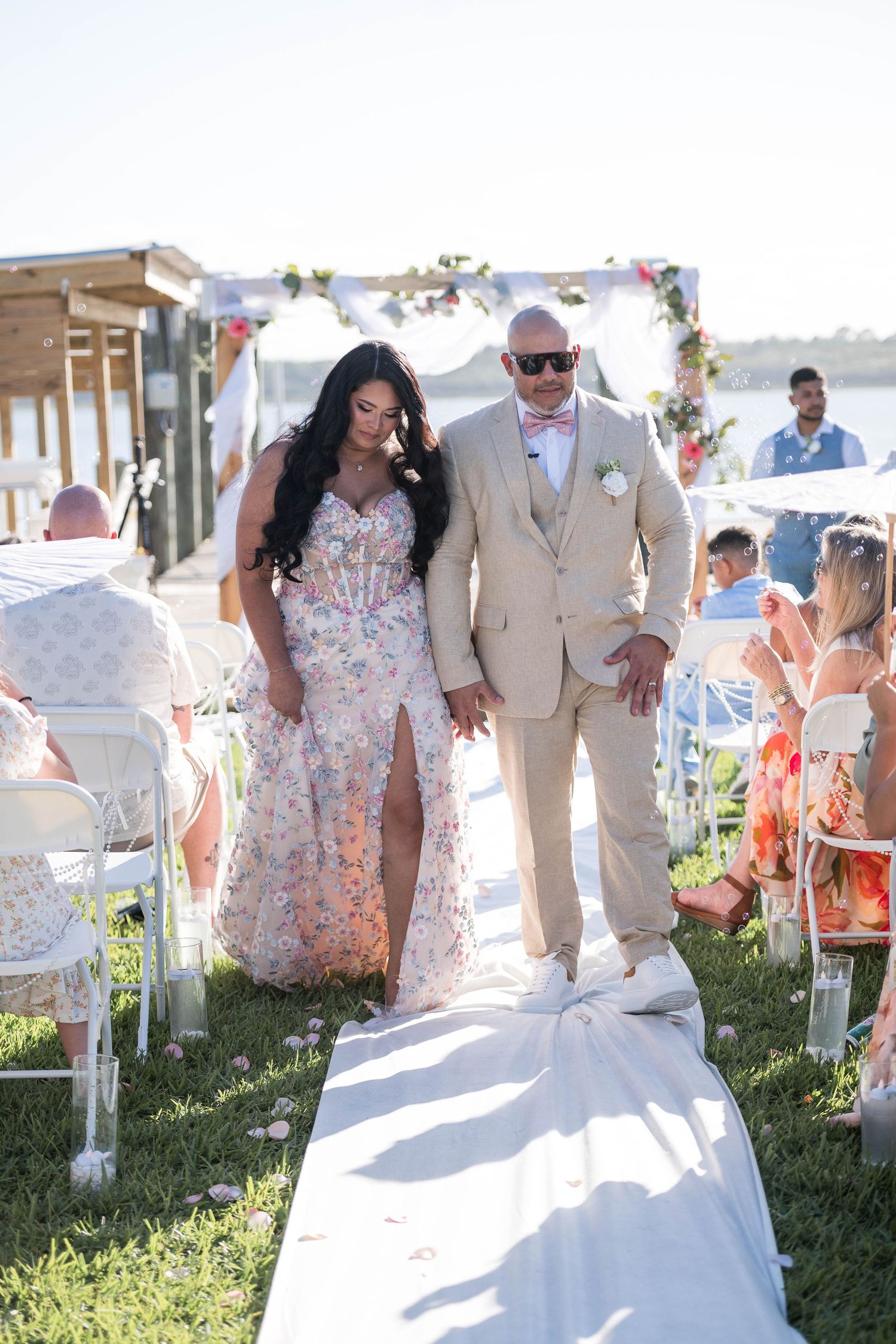Bride and groom walk down outdoor wedding aisle, she in floral gown, he in tan suit with pink bow tie.