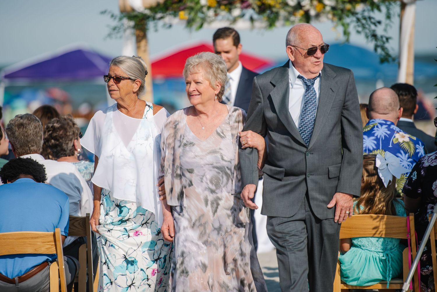 Wedding guests mingling at a beachside ceremony with wooden chairs and floral arrangements.
