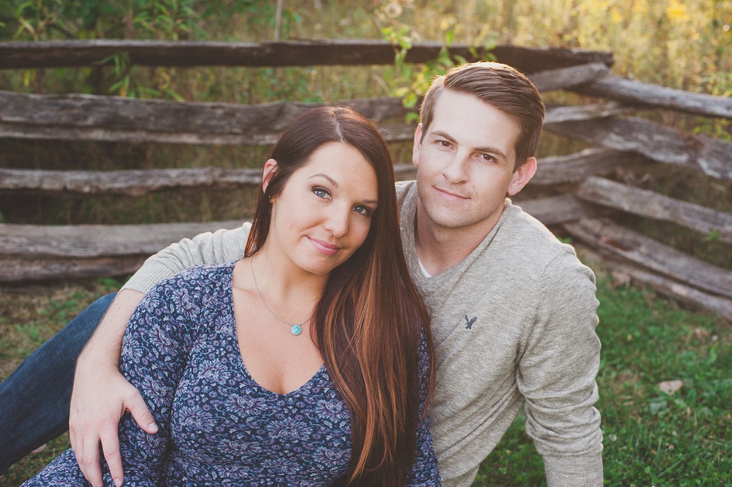 Couple poses casually by wooden split-rail fence during outdoor photo session.